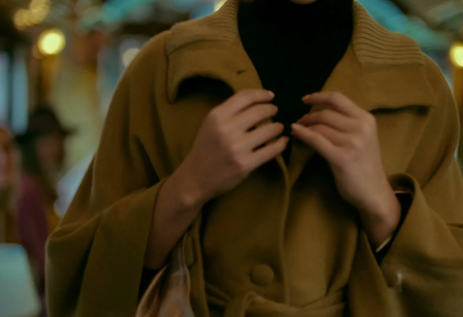 A woman in her 20s, wearing a black turtleneck and a camel coat, buttons her coat while riding a train.
