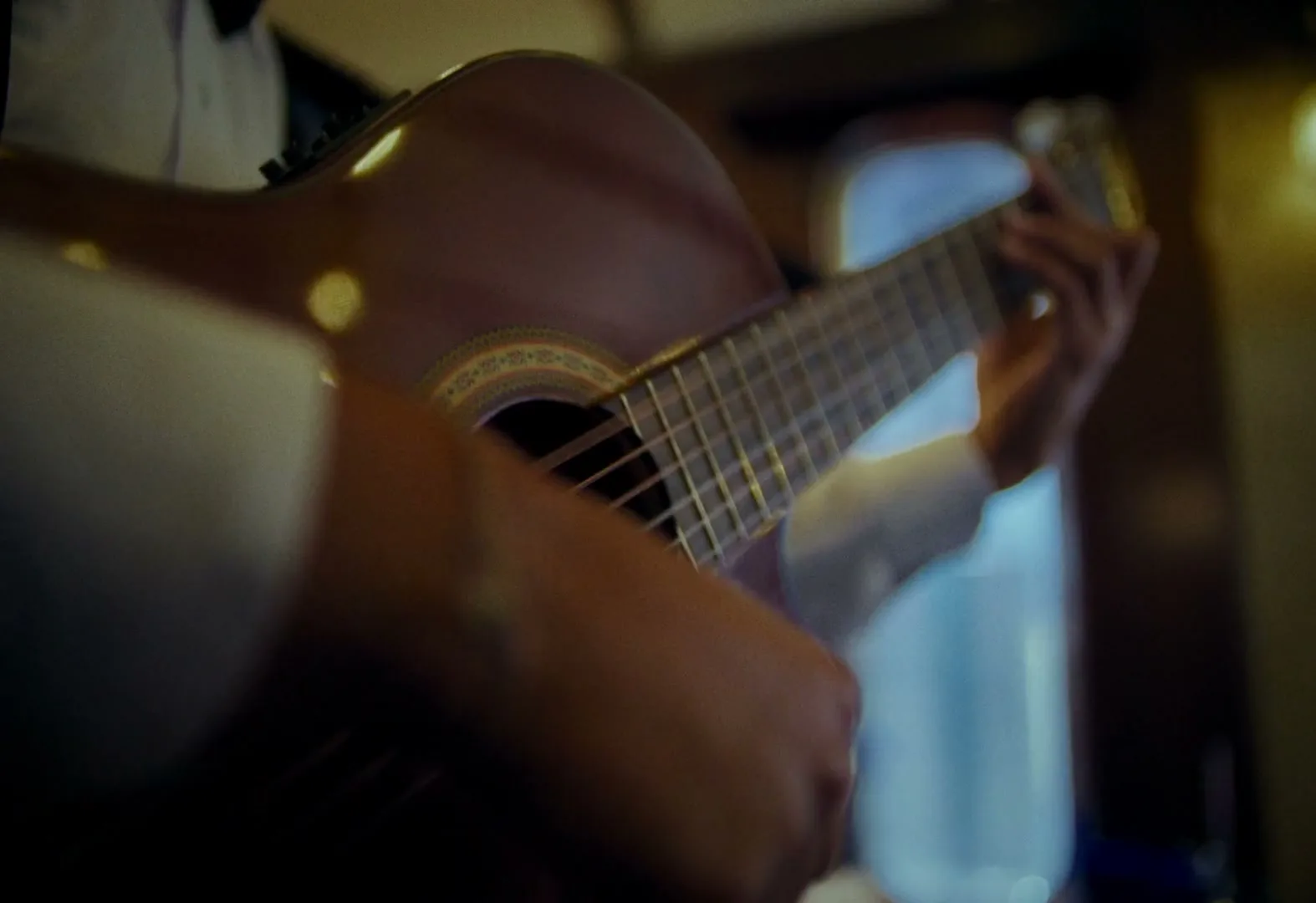 A person in a white shirt plays a classical guitar, strumming the strings with their right hand, inside a dimly lit train car.
