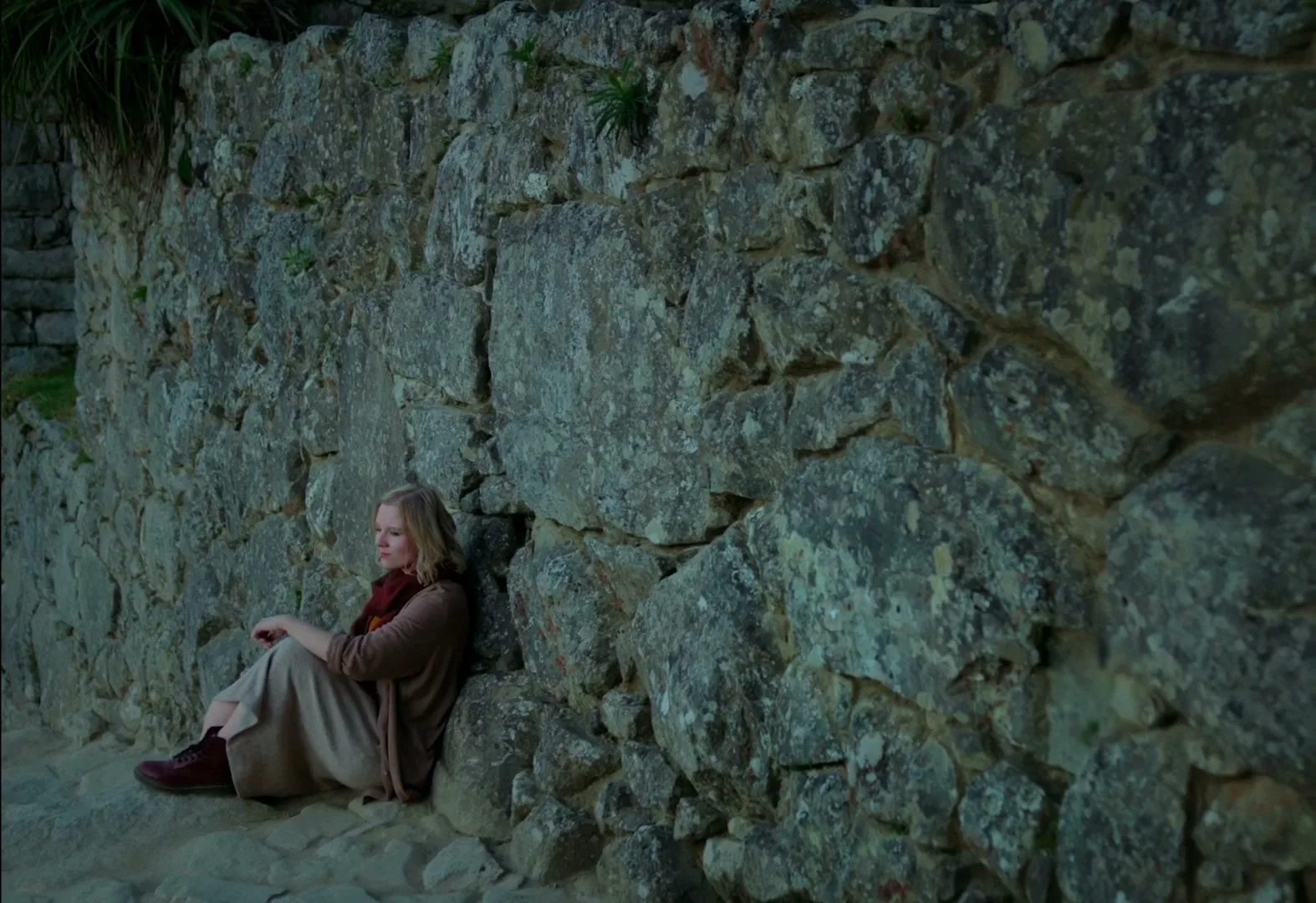 A blonde woman in a brown cardigan and beige skirt sits on the ground, leaning against a stone wall outdoors.
