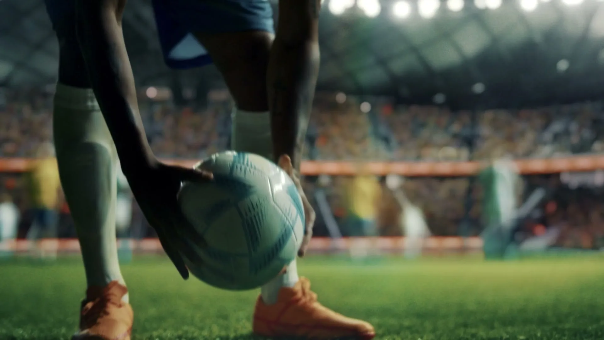 A soccer player in light blue shorts and orange cleats places a white and blue soccer ball on the green grass of a stadium field, with blurred spectators in the background.