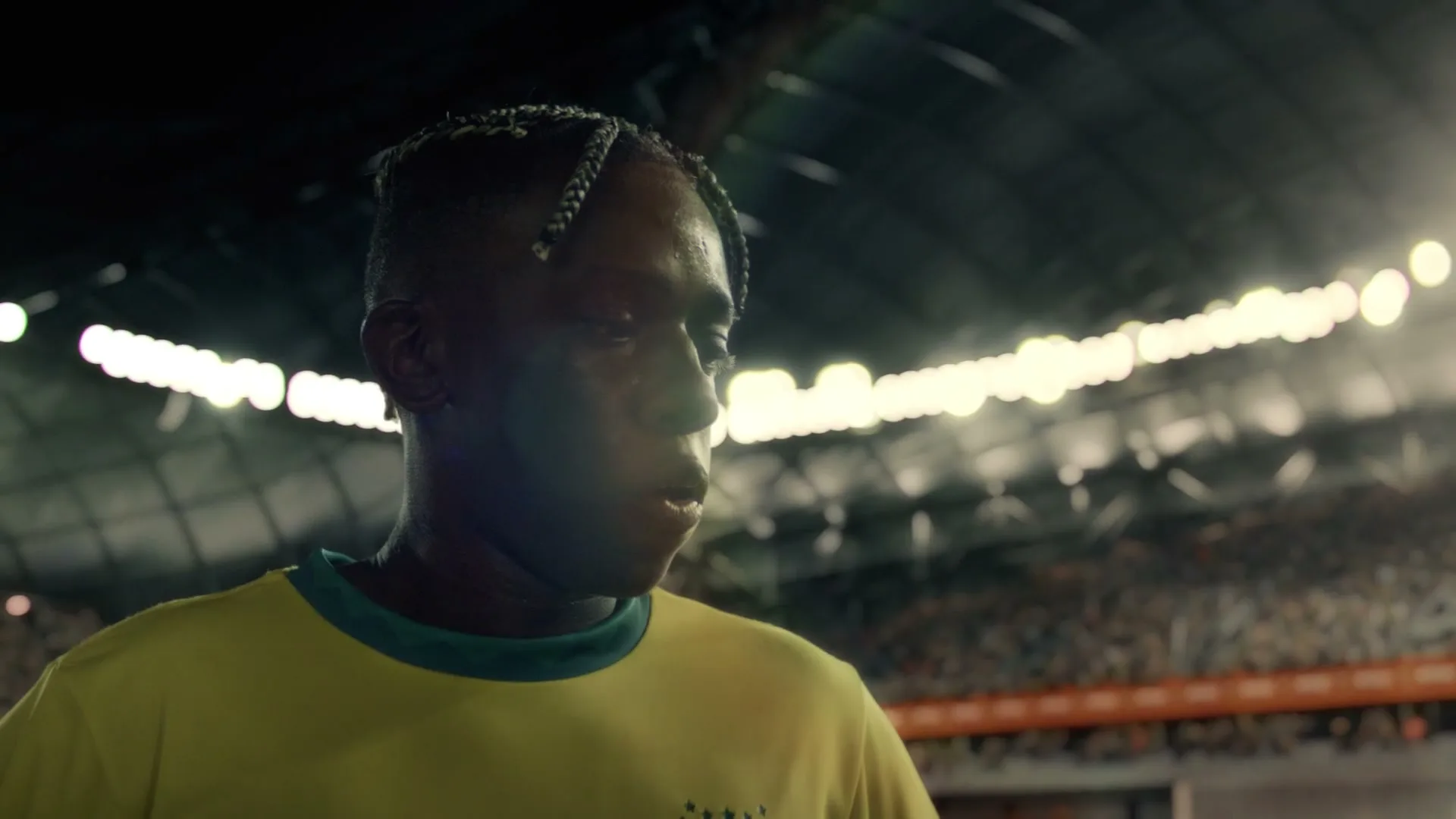 A male soccer player in a yellow jersey looks down, sweating, on a brightly lit stadium field at night.