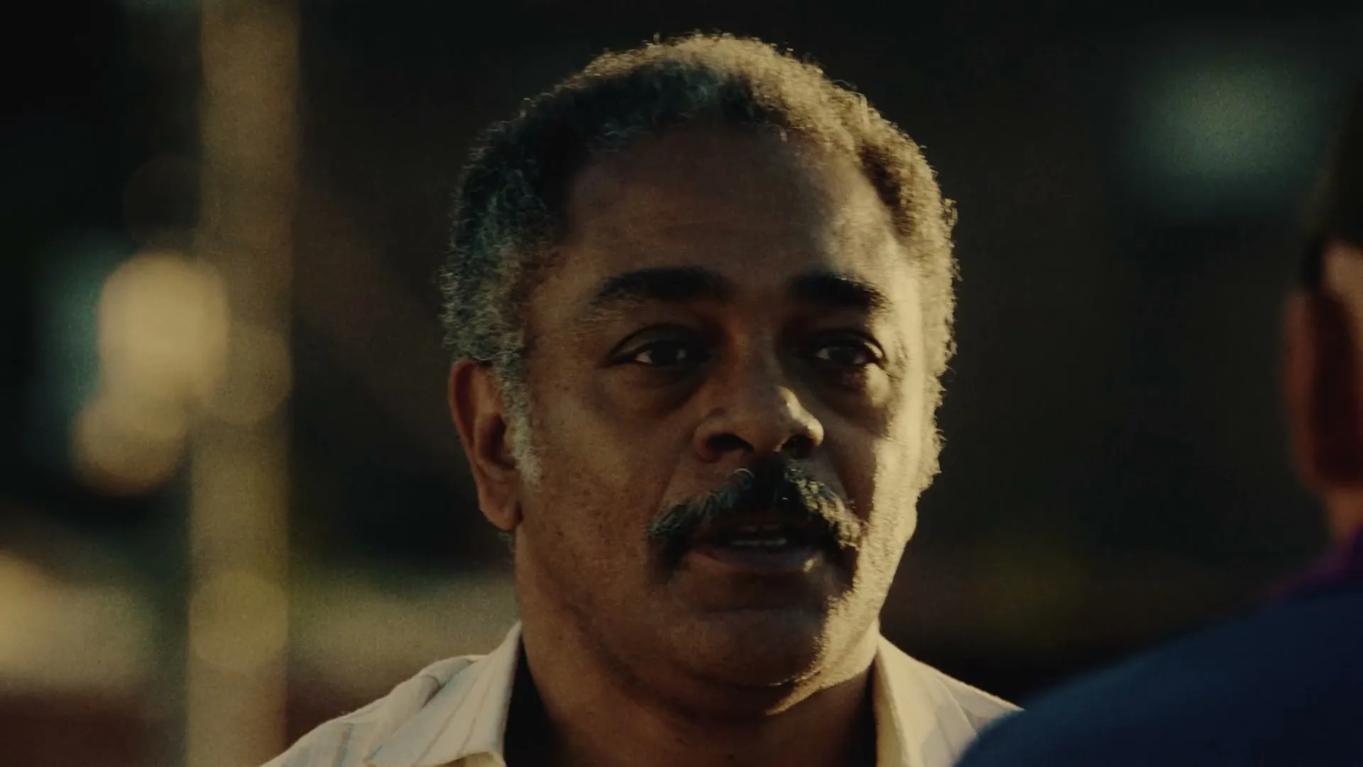 A middle-aged man with a mustache looks intently at someone off-screen, his expression serious, in an outdoor setting at dusk.