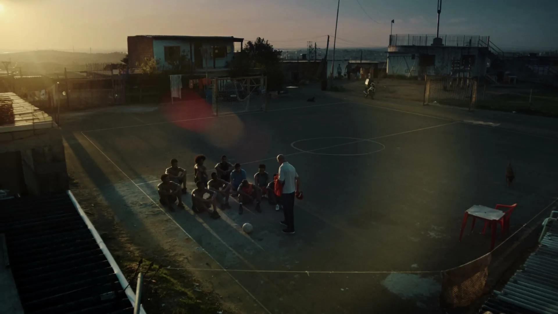 A group of young boys and a man are gathered on a dusty soccer field, with buildings and a sunset in the background.