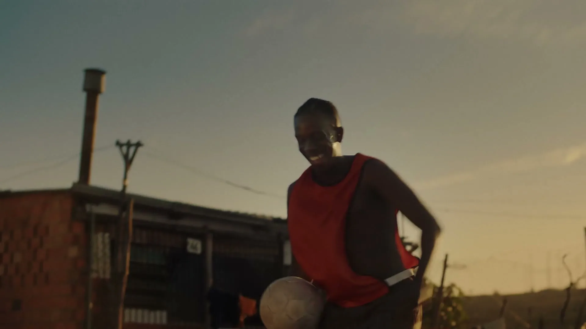 A young man in a red vest smiles and juggles a soccer ball in a dusty, outdoor setting at golden hour.
