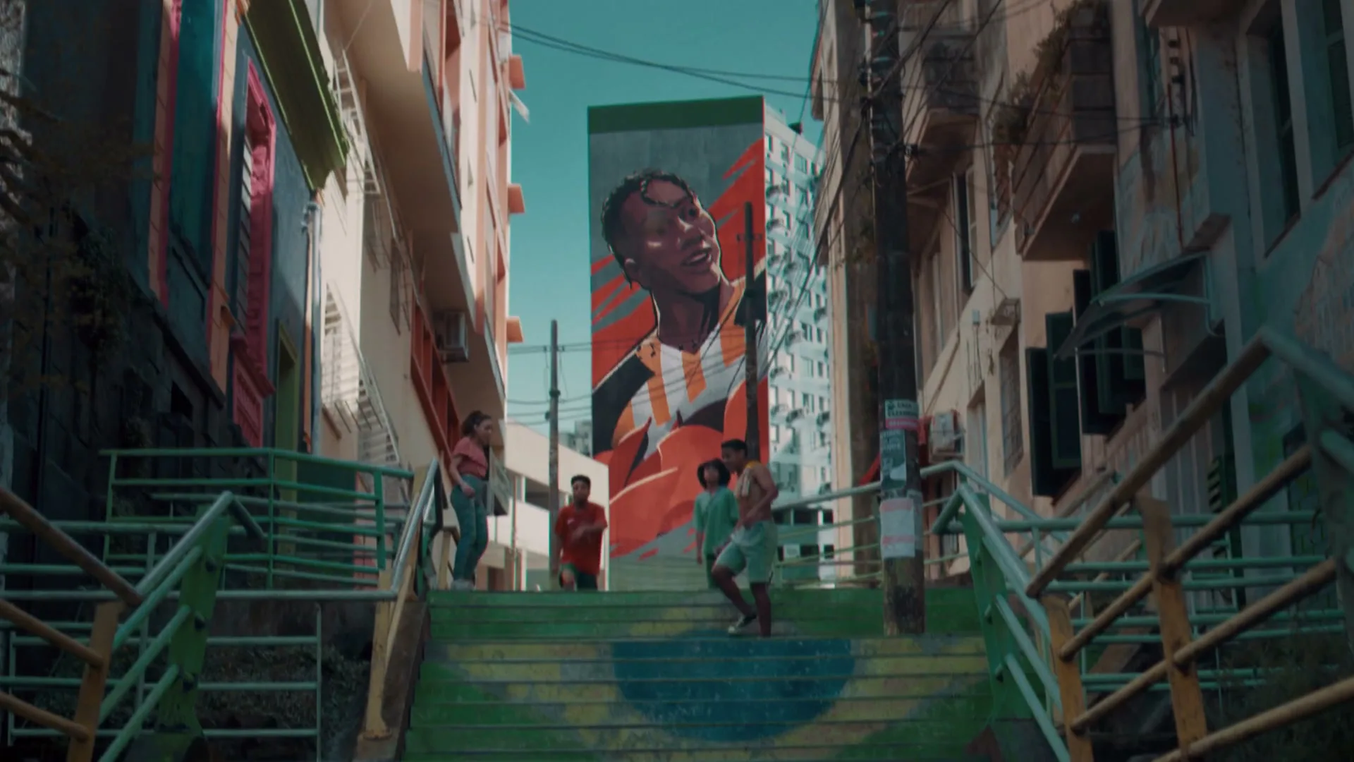 A group of people walk down a colorful staircase in a narrow alleyway, with a large mural of a smiling man on a building in the background.