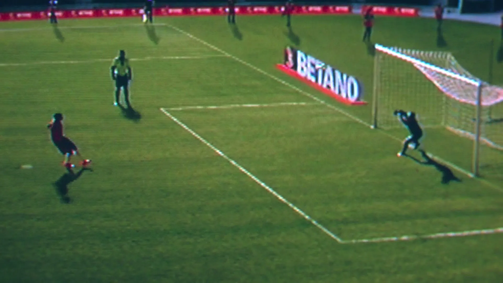 A soccer player in a red jersey kicks a penalty shot towards the goal, defended by a goalie in a black jersey, on a green field during the day.