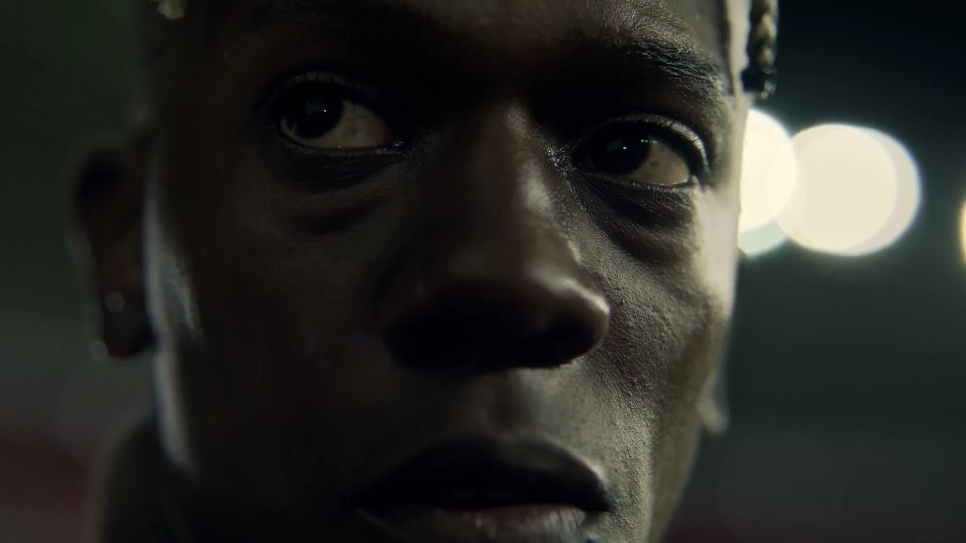 A young Black man with braided hair looks up with a serious expression, illuminated by soft light, at night.