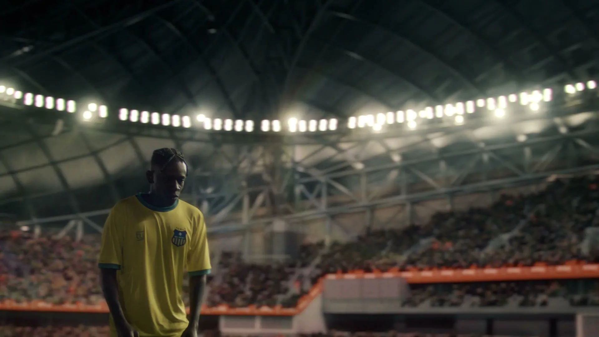 A male soccer player in a yellow jersey stands on a field in a large, brightly lit stadium with a crowd in the background.