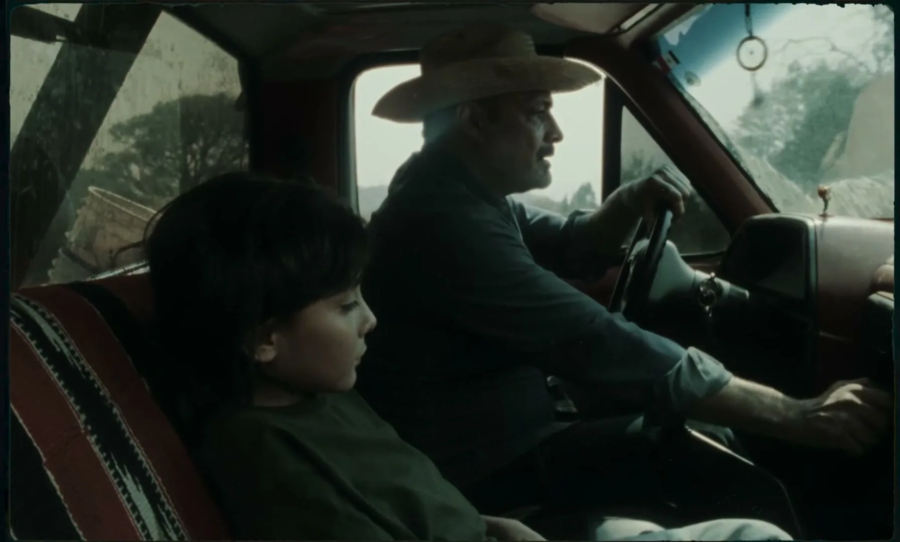 A young child with dark hair sits in the passenger seat of a truck, looking forward, while an older man in a cowboy hat drives, both appearing somber.