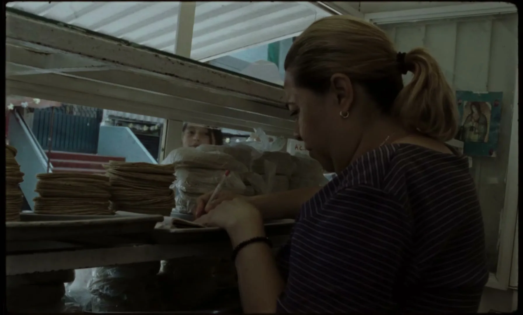 A woman with blonde hair in a ponytail writes in a notebook behind a counter with stacks of tortillas, while a child's face appears in the background.