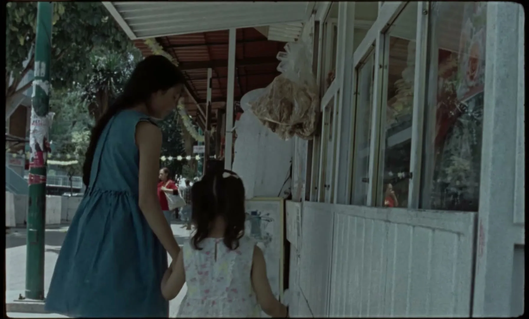 A young woman in a blue dress and a little girl in a white dress walk hand-in-hand past a street vendor stall on a sunny day.