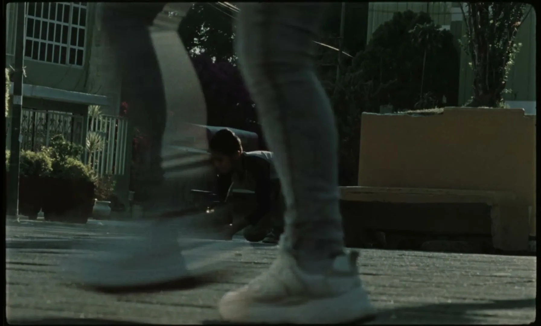 A young boy kneels on a street, picking up a radio, as people run past him in a residential area during the day.