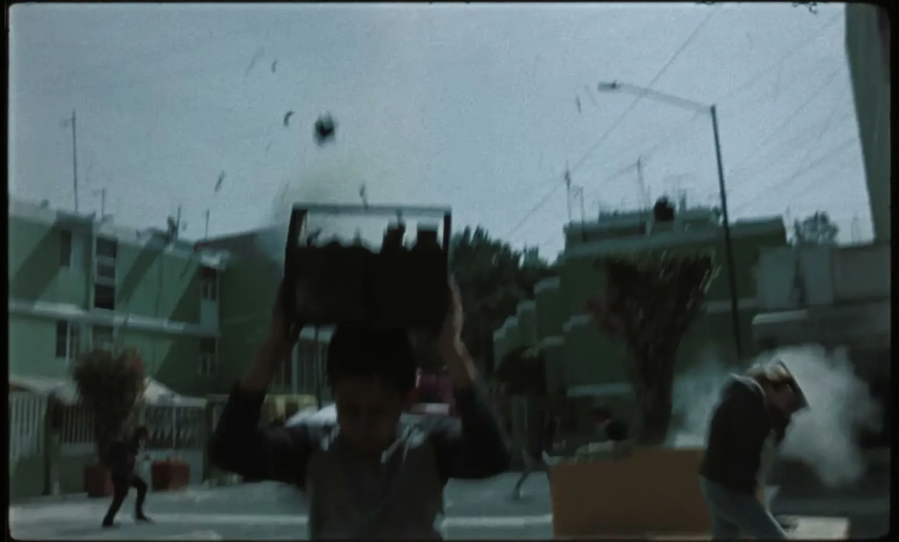 A young boy walks down a street carrying a makeshift cable car on his head as debris falls around him.