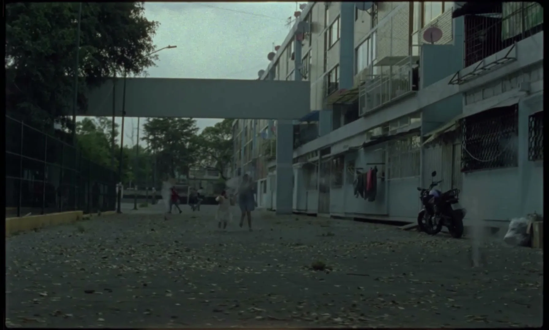 A group of children run and play in a dusty, debris-strewn alleyway between apartment buildings, as a person runs past the camera.