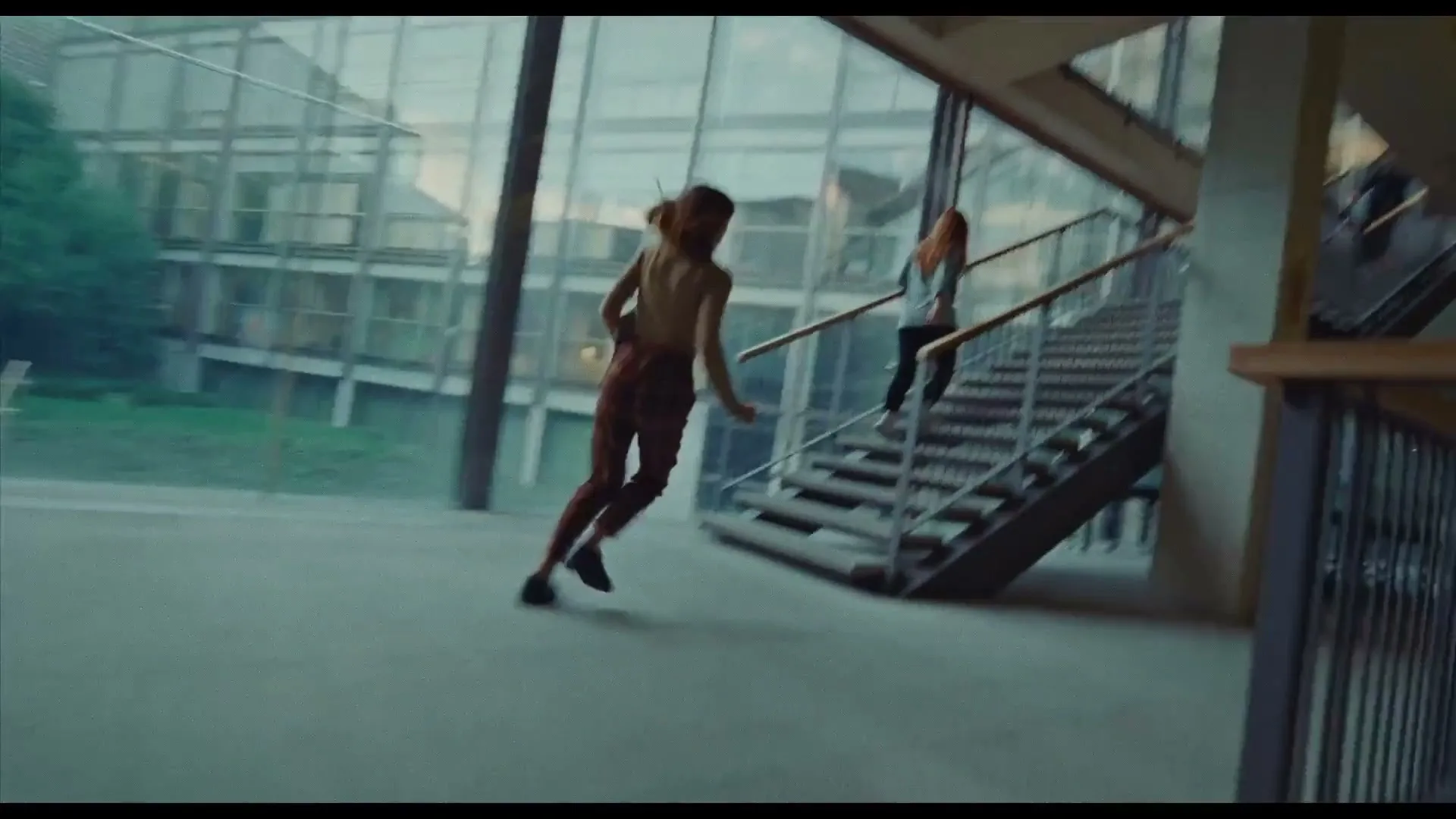 A young woman in a tank top and pants runs towards and up a modern staircase, while another woman walks up the stairs in a bright, airy building.