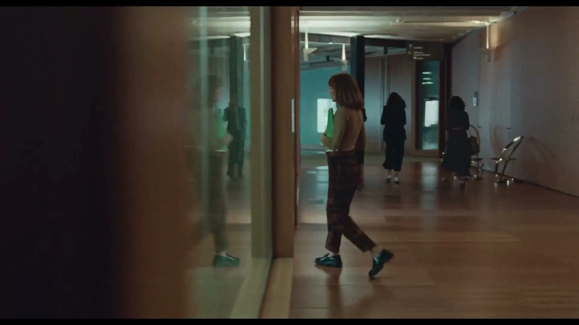 A young woman with brown hair, wearing a green top and plaid pants, walks down a modern hallway with glass walls and other people in the background.