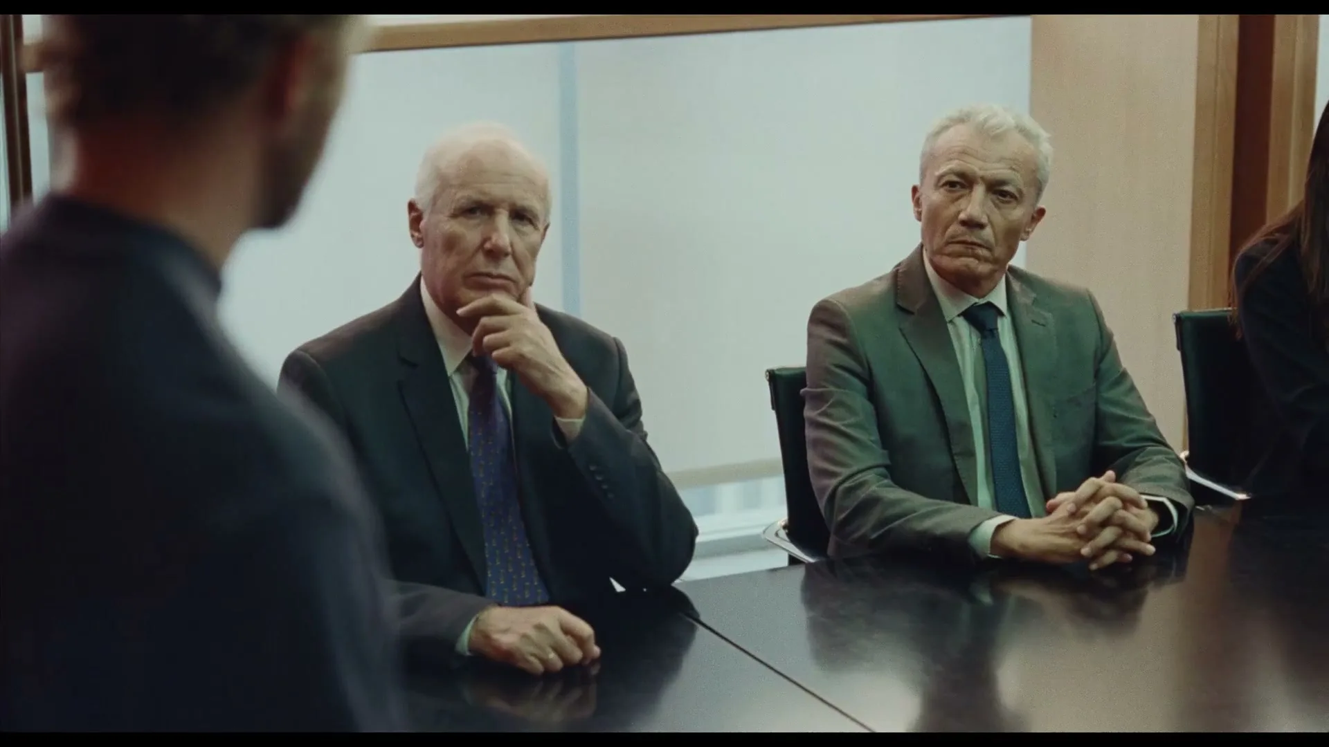 Two older men in suits sit at a conference table, listening intently to someone off-screen in a modern office.
