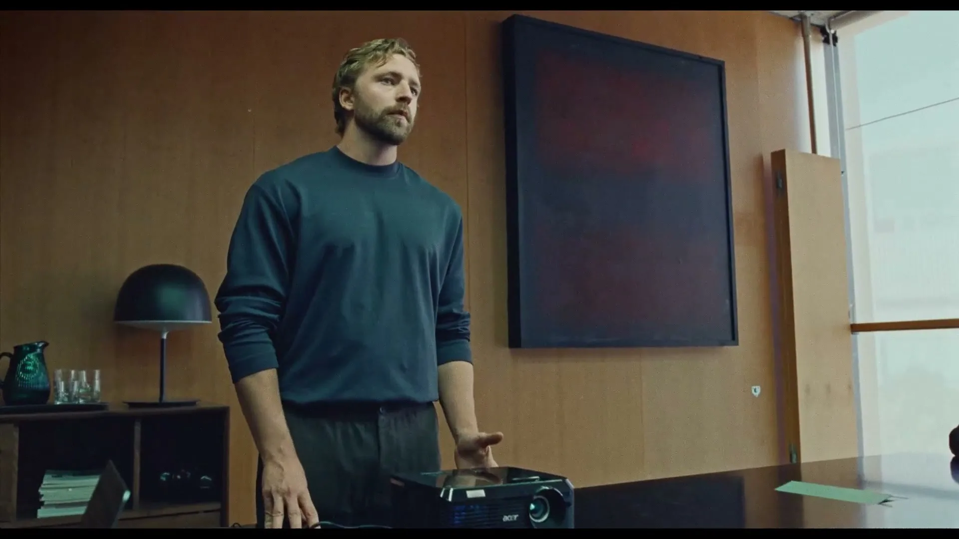 A man with blonde hair and a beard stands next to a projector on a conference table, gesturing with his hands in a modern office.