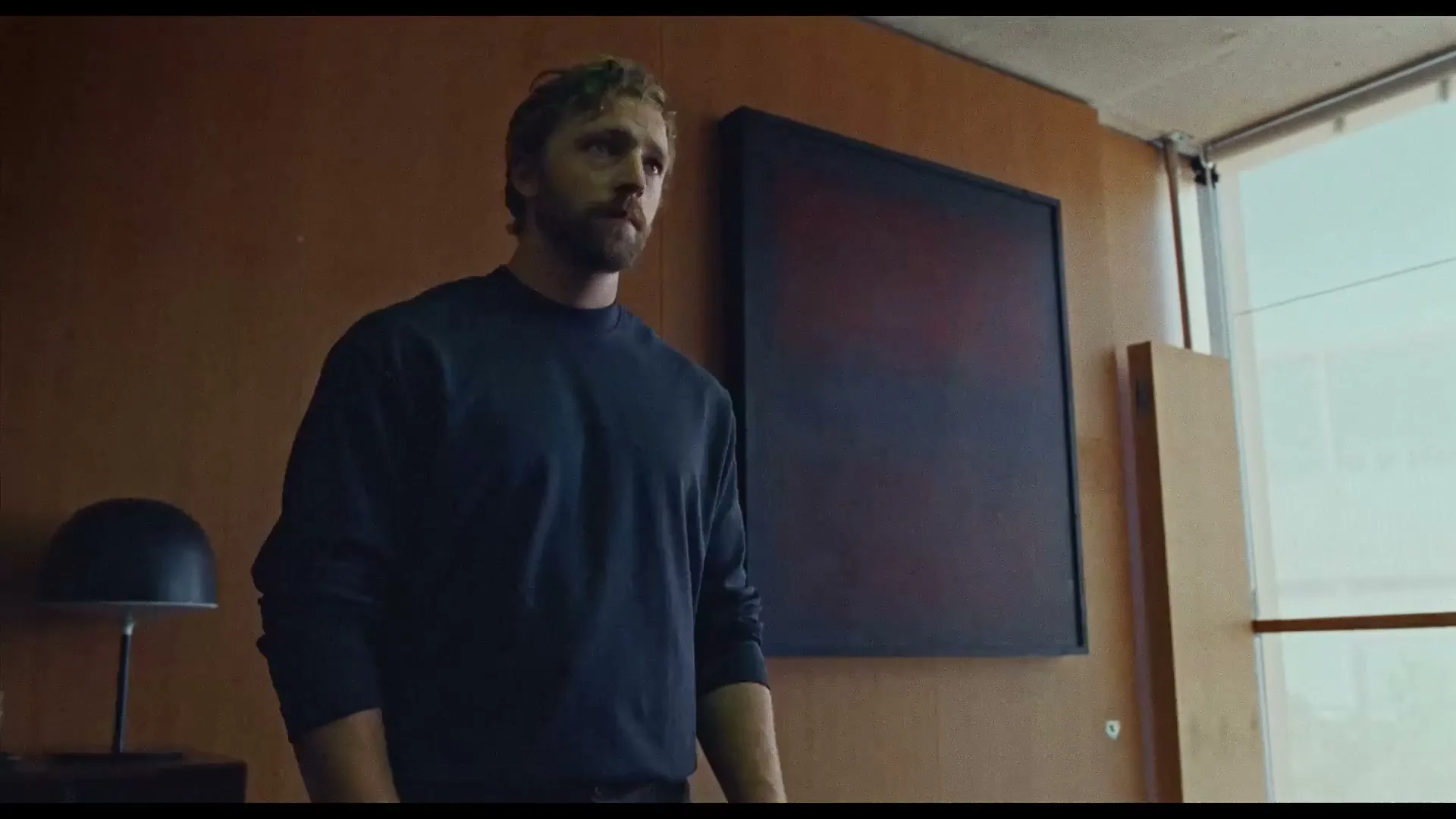 A man with a beard and curly hair stands in a modern room with wood paneling and a dark painting, looking down thoughtfully.