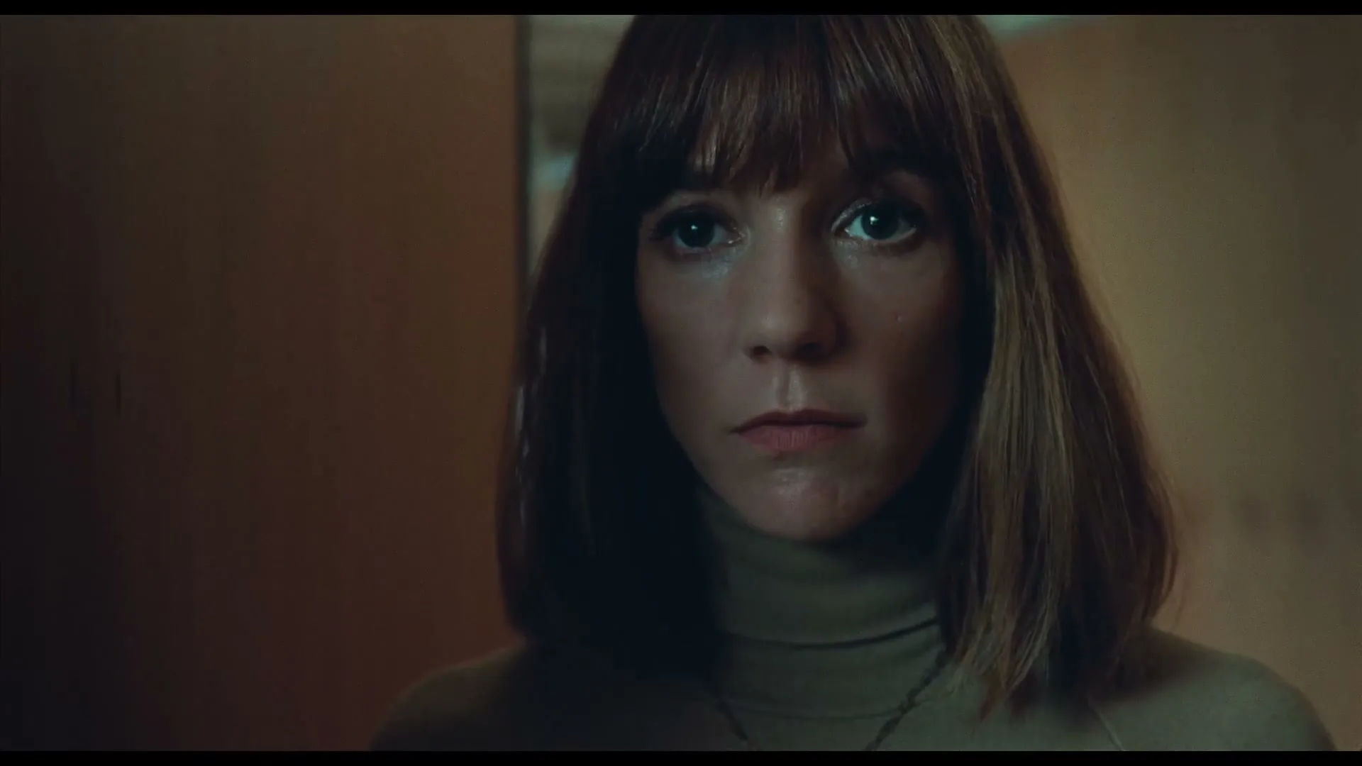 A woman with bangs and a turtleneck looks directly at the camera with a serious expression in a dimly lit room.