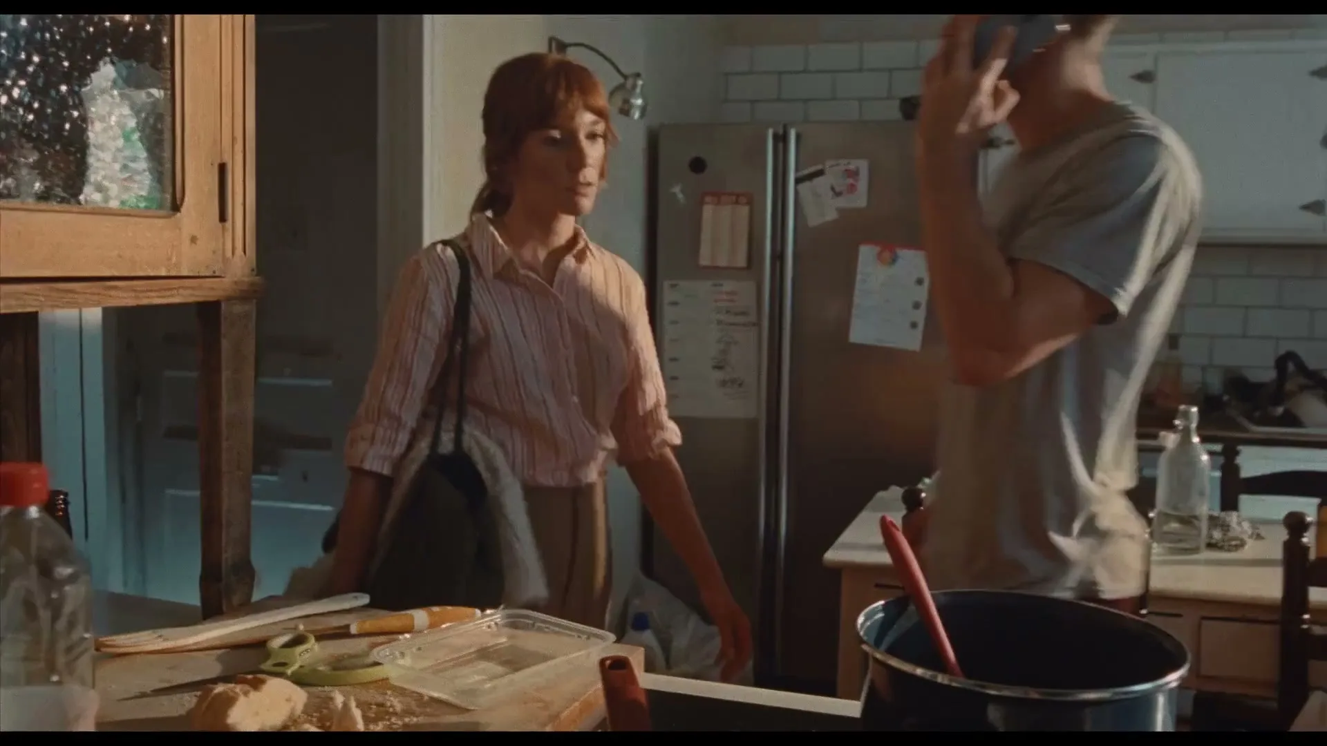 A woman with reddish-brown hair and a striped shirt stands in a kitchen, looking around as a man walks past her in the background.