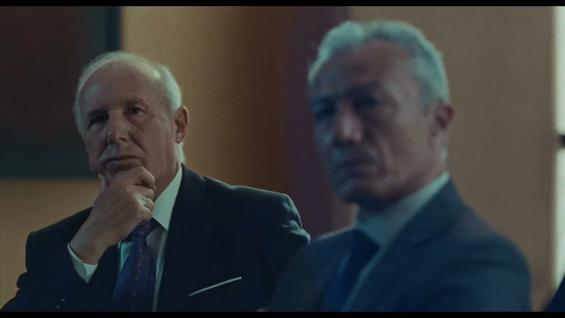 Two older men in suits are seated in an interior setting, with one man in the foreground looking thoughtful and the other man in the background out of focus.