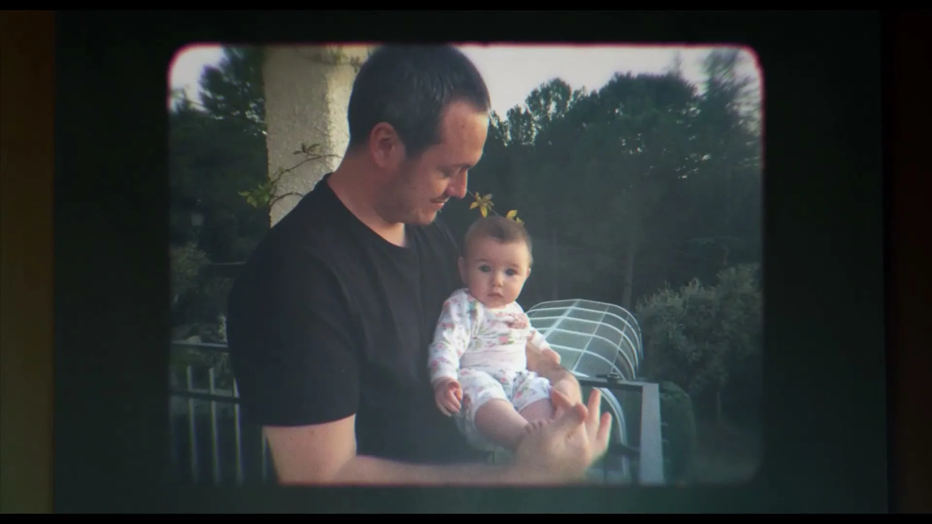 A man in a black t-shirt holds a baby in a white onesie, looking down at the baby with a gentle smile, outdoors on a patio.