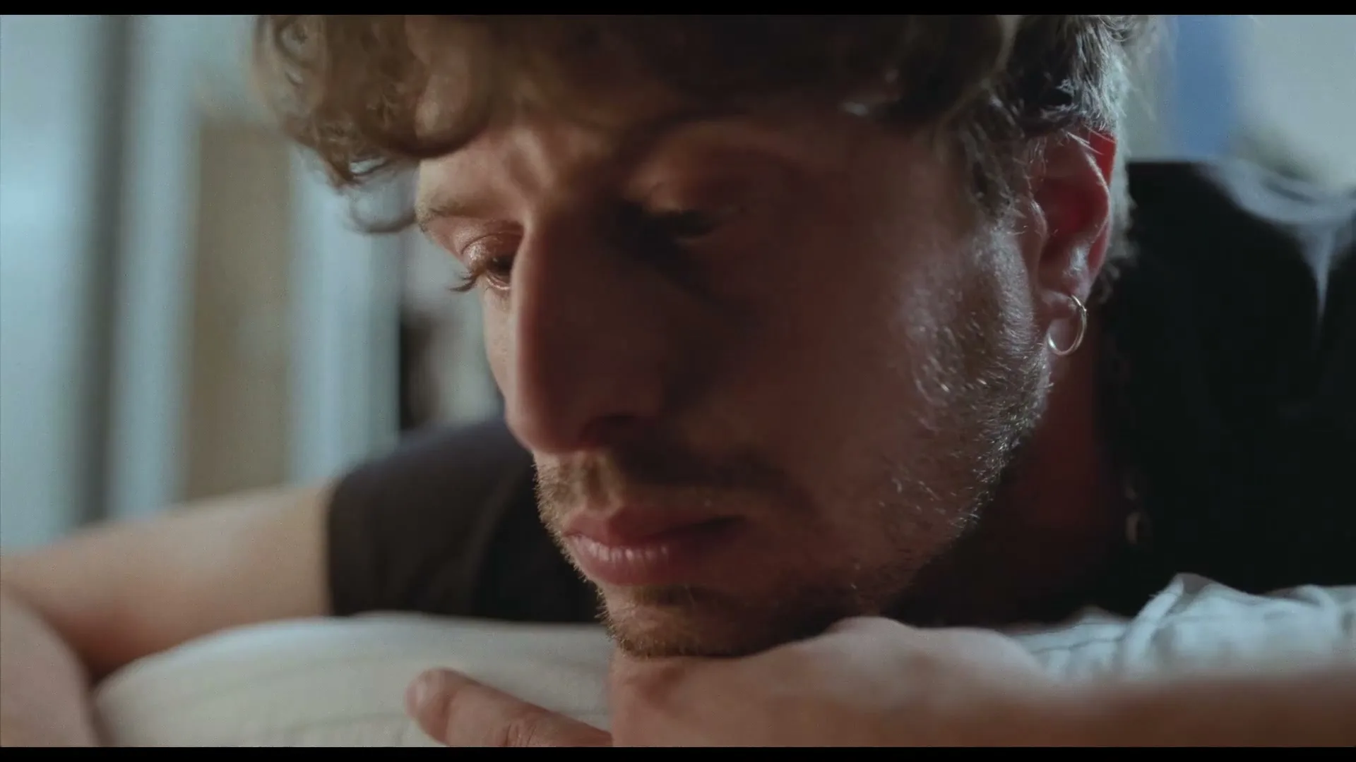 A man with curly hair and a beard lies on a pillow, looking down with a pensive expression in an interior setting.
