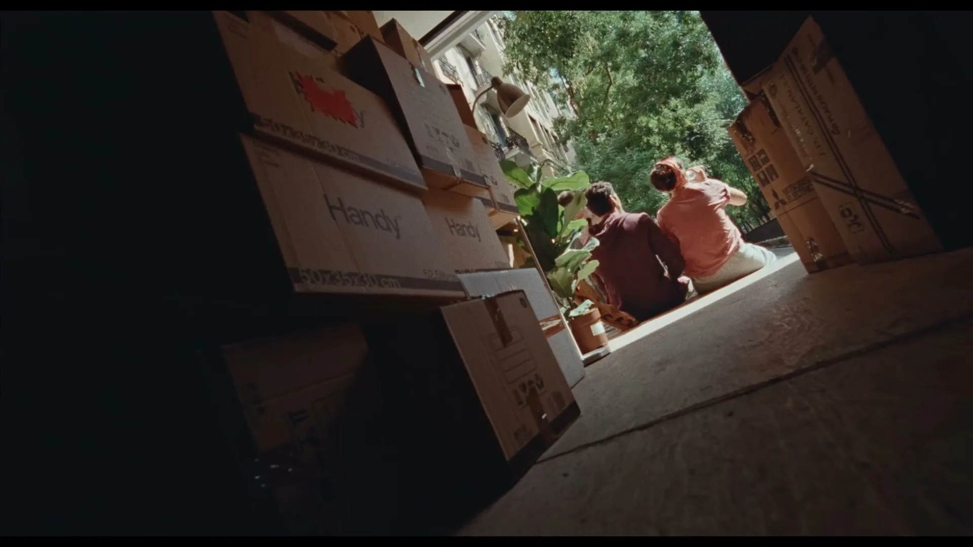 Three men are seen from inside a moving truck, sitting on the ground and talking, surrounded by moving boxes and plants, on a sunny day.