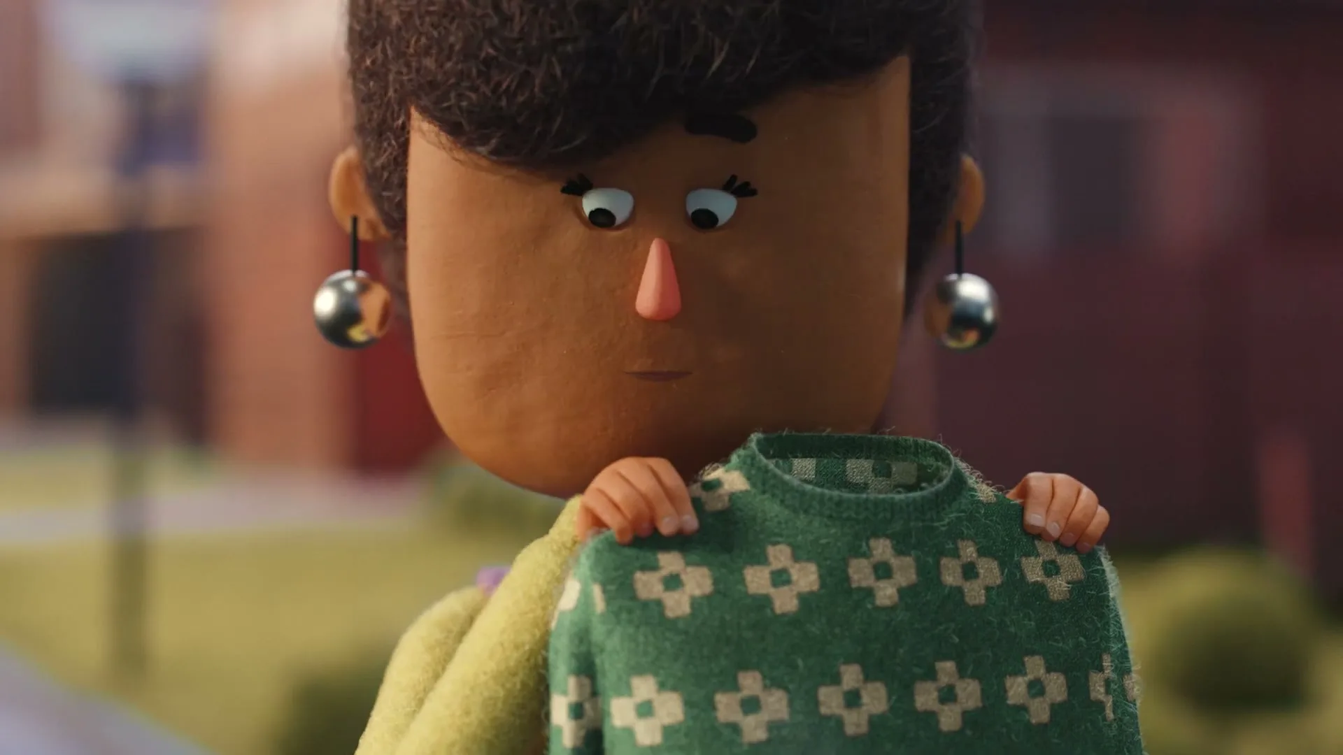 A claymation-style woman with dark hair and large earrings holds up a green patterned sweater, looking at it thoughtfully outdoors.