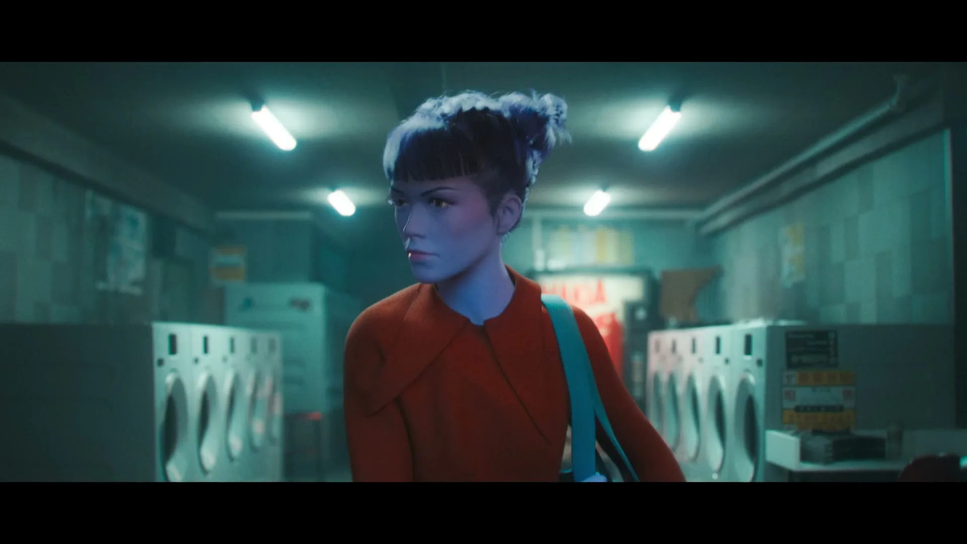 A woman with blue skin and hair, wearing an orange jacket, turns to face the camera in a dimly lit laundromat.