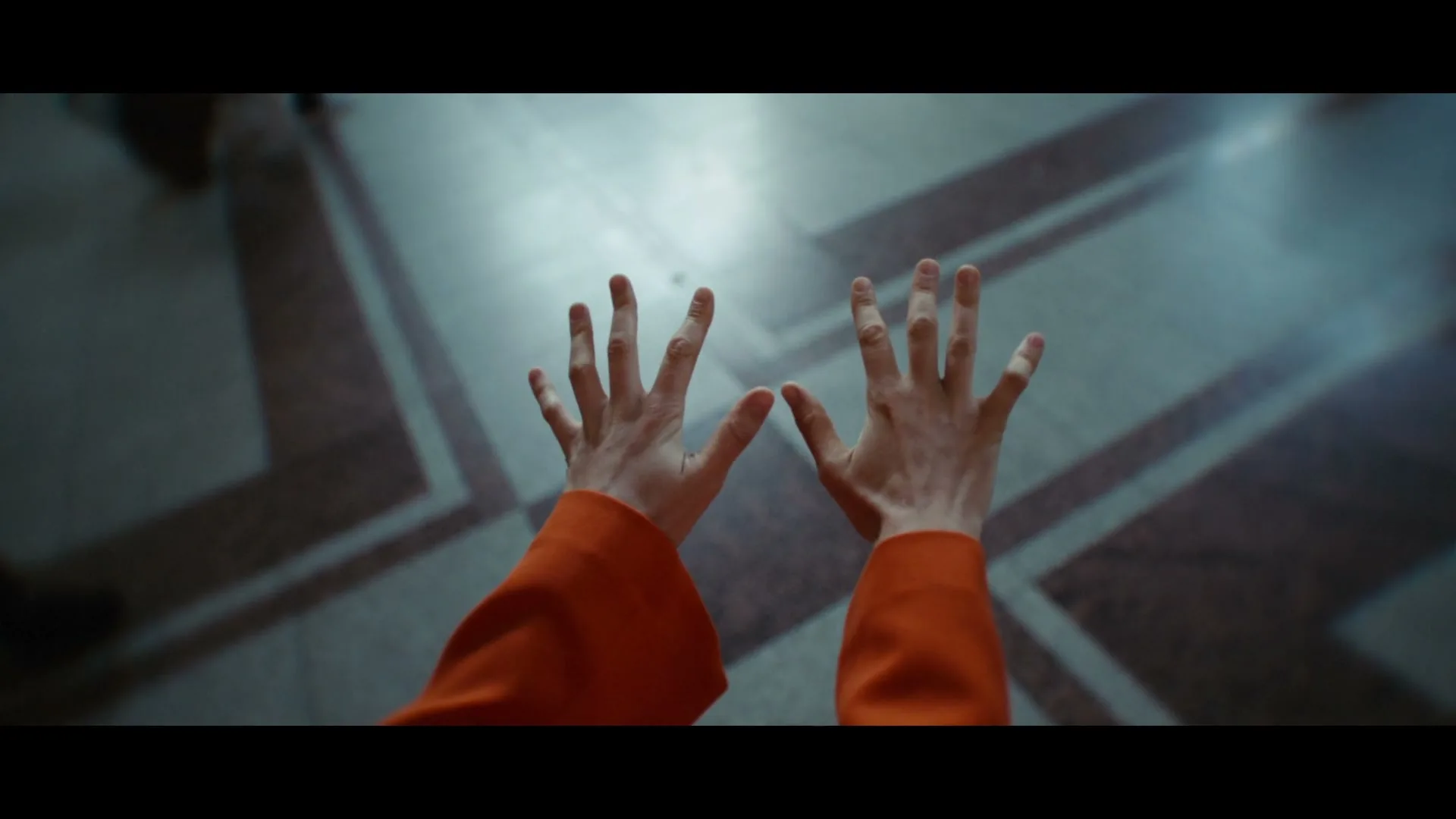 A person in an orange top raises their hands towards the camera, as another person walks past them on a patterned floor.