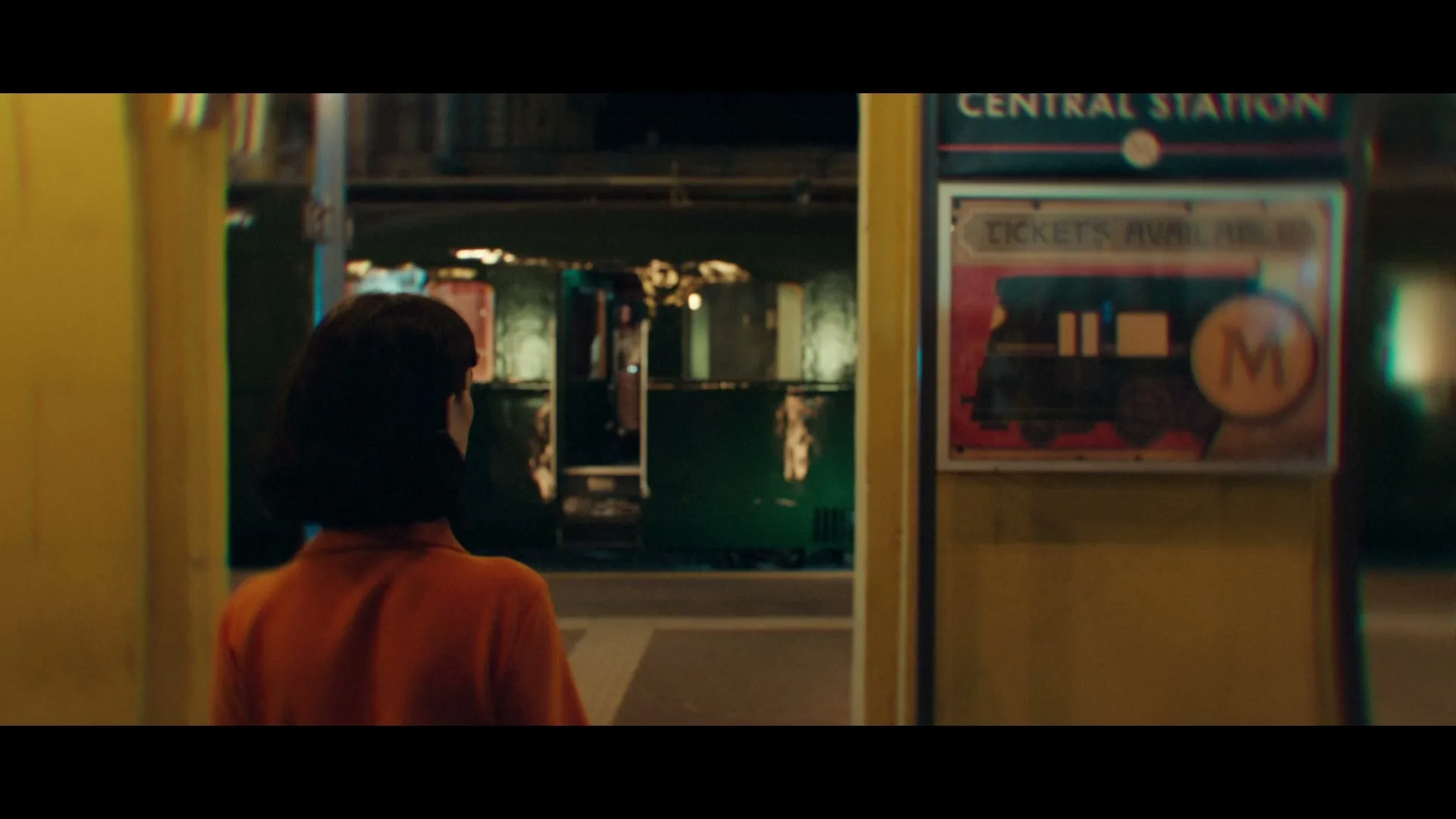 A woman with dark hair in an orange top stands on a train platform at night, looking towards a train and a "Central Station" sign.