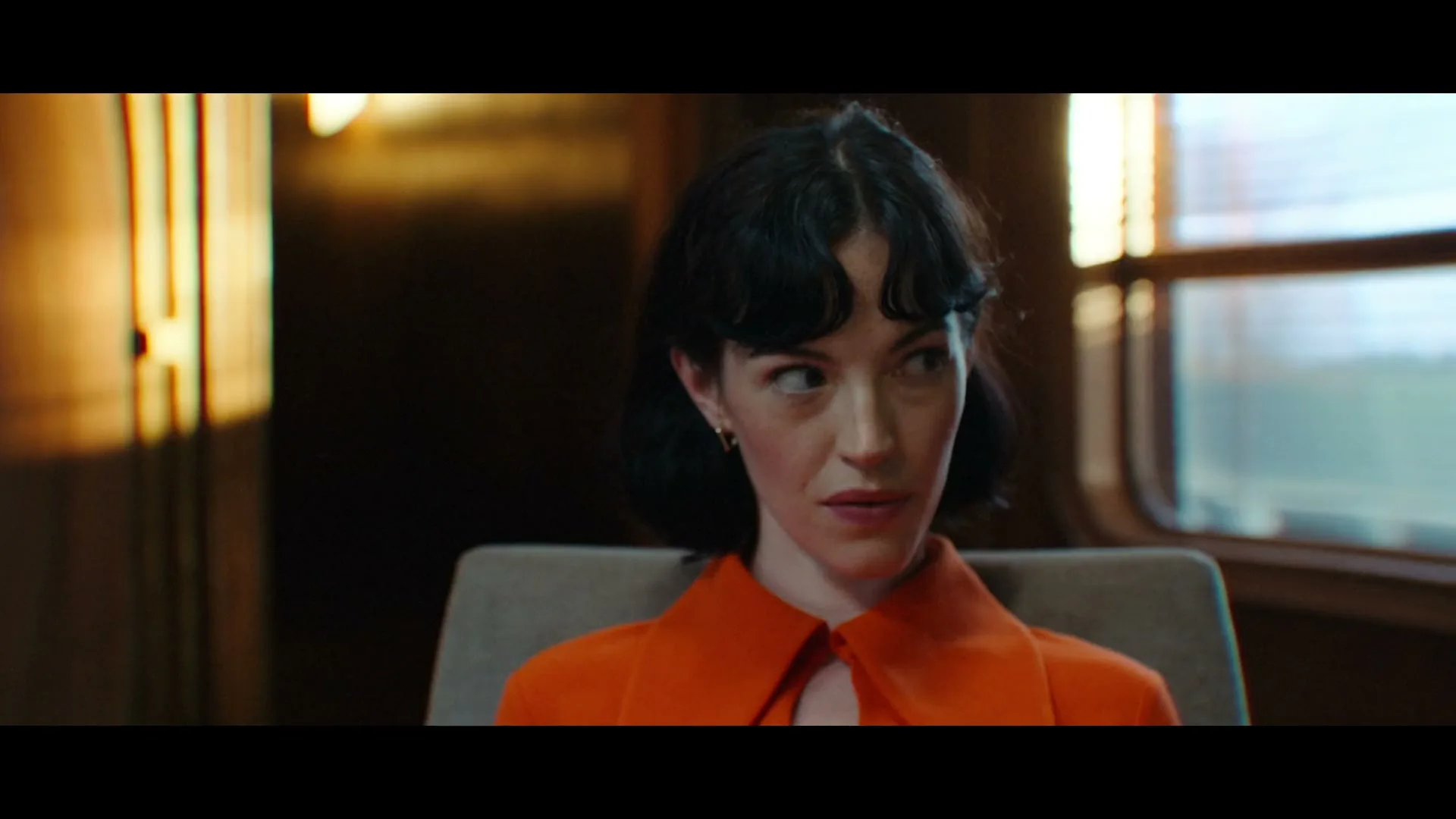 A young woman with dark hair and an orange top sits in a train, looking around with a curious expression.