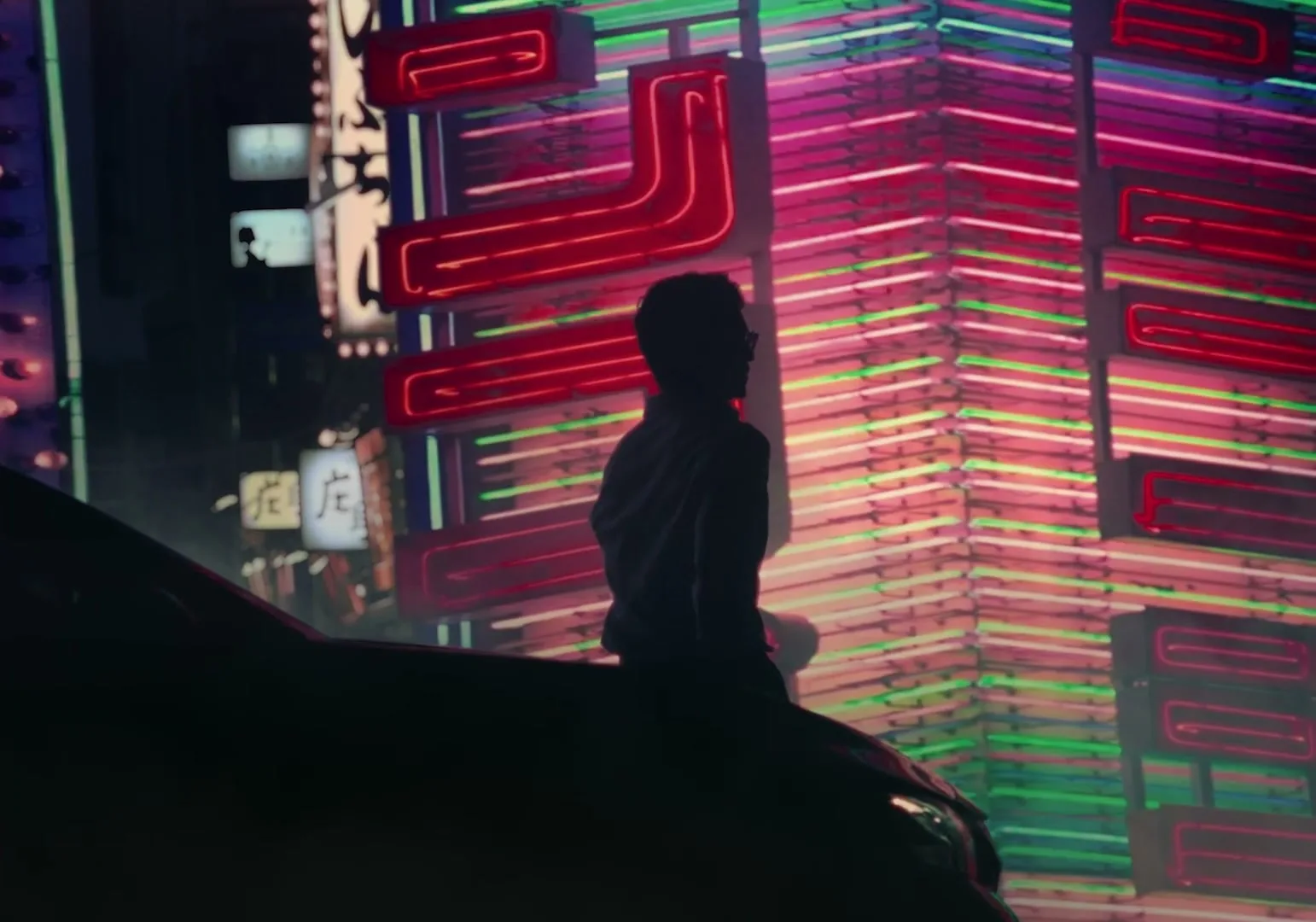 A man in silhouette sits on a car hood, looking at a building covered in bright neon signs at night in a city.