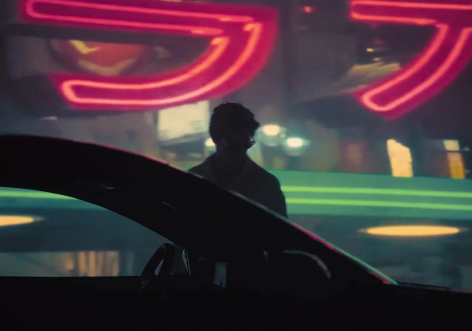 A man with dark, curly hair stands in front of a bright pink neon sign, partially obscured by the silhouette of a car's roof and windshield.