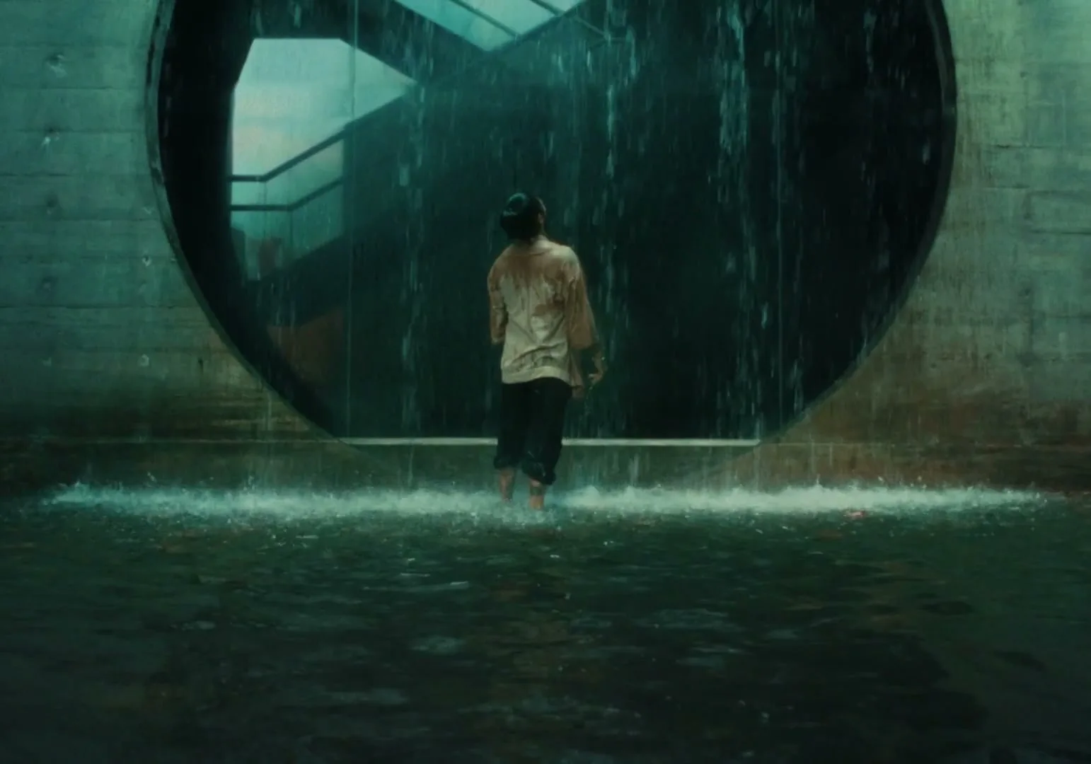A man in a light shirt and dark pants stands barefoot in shallow water, facing a large circular opening with water cascading down in an interior setting.