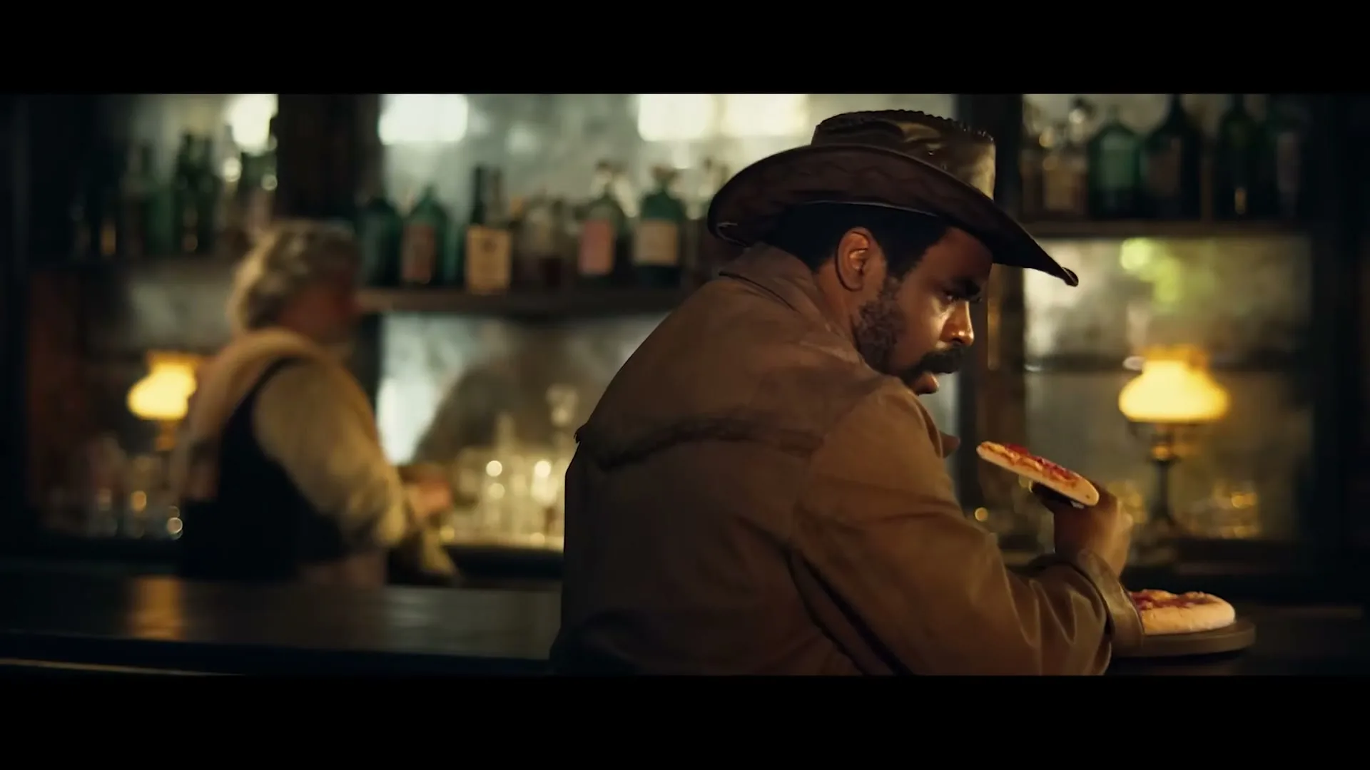 A man in a cowboy hat and jacket sits at a bar, eating a slice of pizza, then turns to look over his shoulder in a dimly lit saloon.