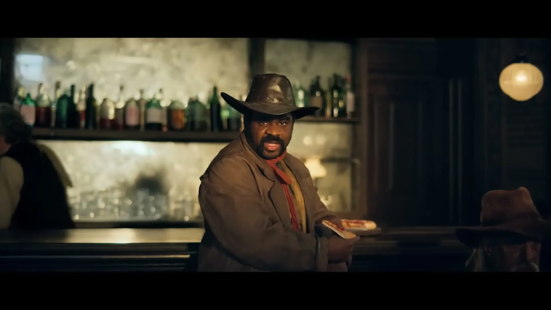 A man in a cowboy hat and duster coat holds a slice of pizza, looking intensely at the camera in a dimly lit saloon.