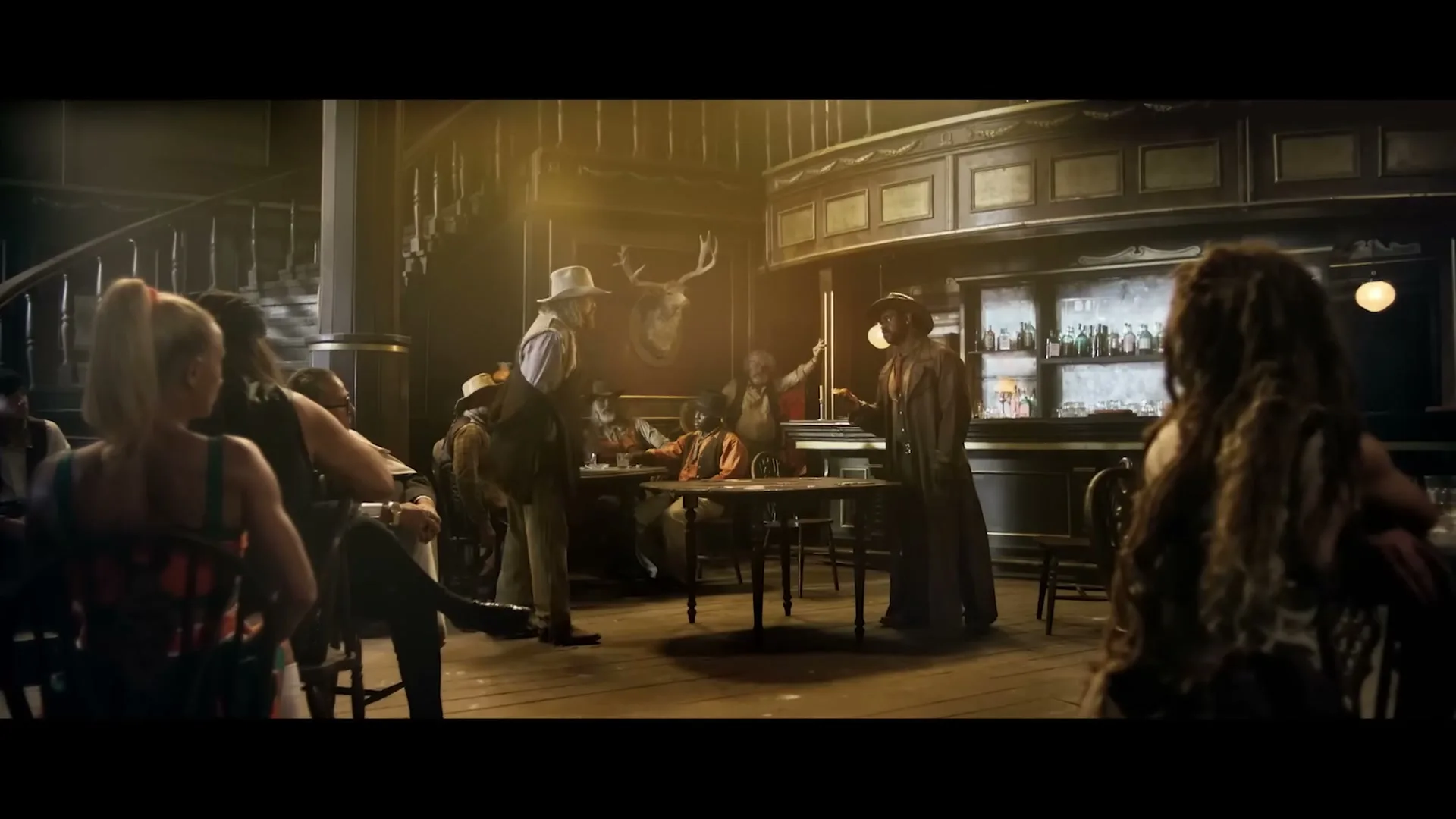 In a dimly lit saloon, a man in a cowboy hat kicks his leg onto a table, facing another man in a long coat, as other patrons watch.