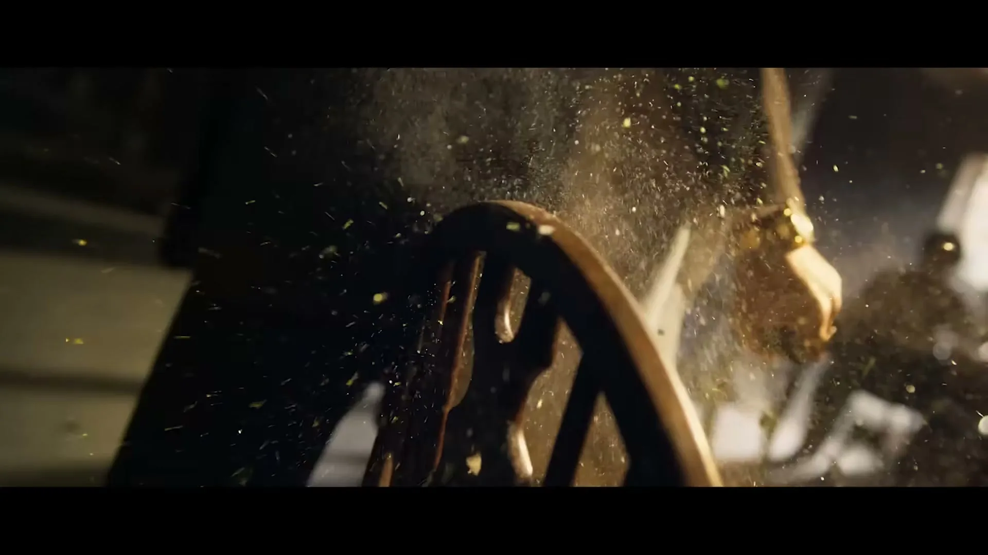 A man in a suit with a gold watch slams his hand on a wooden chair, causing green spices to fly into the air in a dimly lit room.