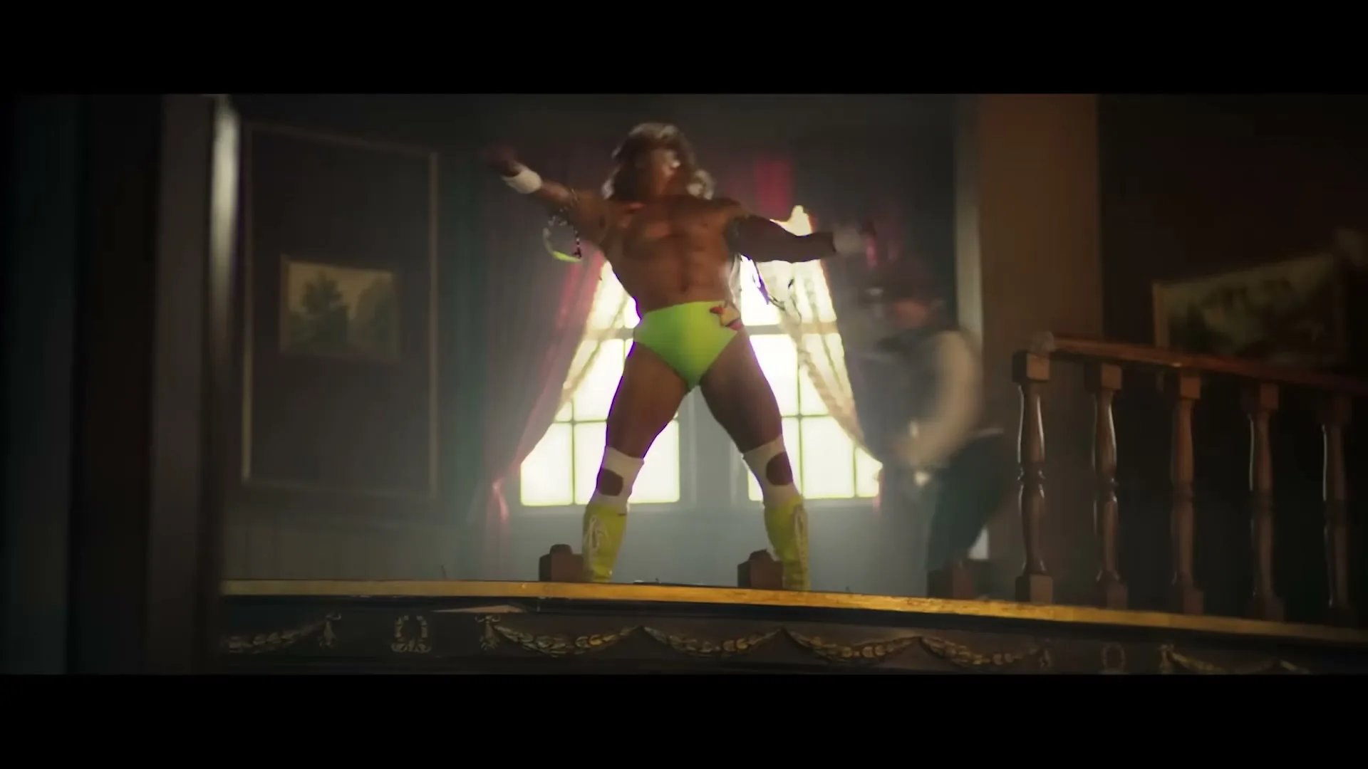 A muscular male wrestler in a neon green speedo and yellow boots flexes on a balcony as two men in cowboy attire approach him in a dimly lit saloon.