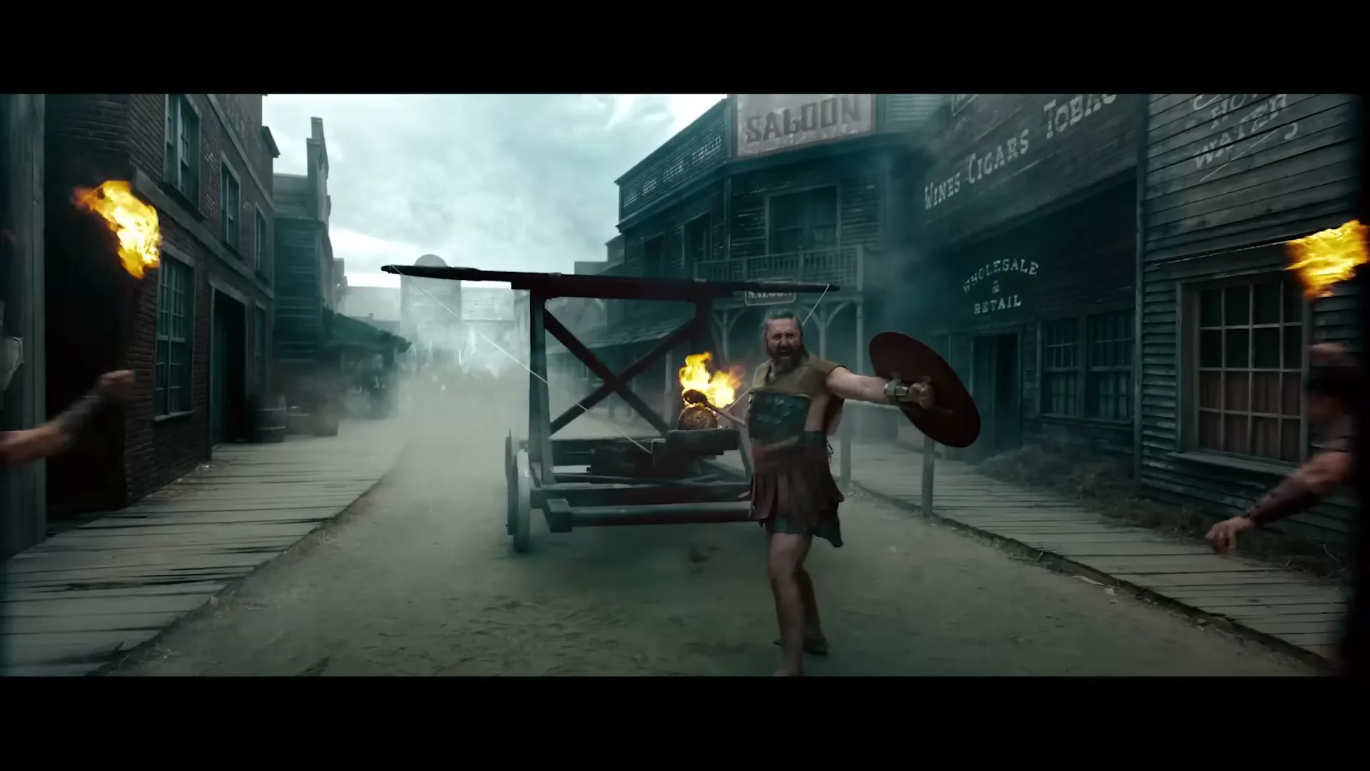 A group of warriors in ancient attire run through a dusty old western town, carrying torches and shields, as a catapult with a flaming pizza on it rolls down the street.