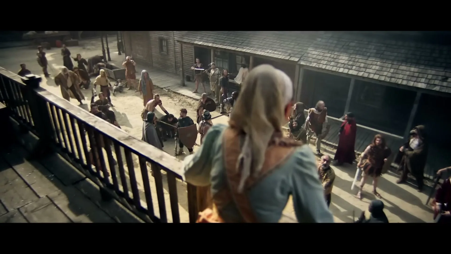 A woman in period clothing looks down from a balcony at a crowd of people in a rustic town square, some holding weapons.