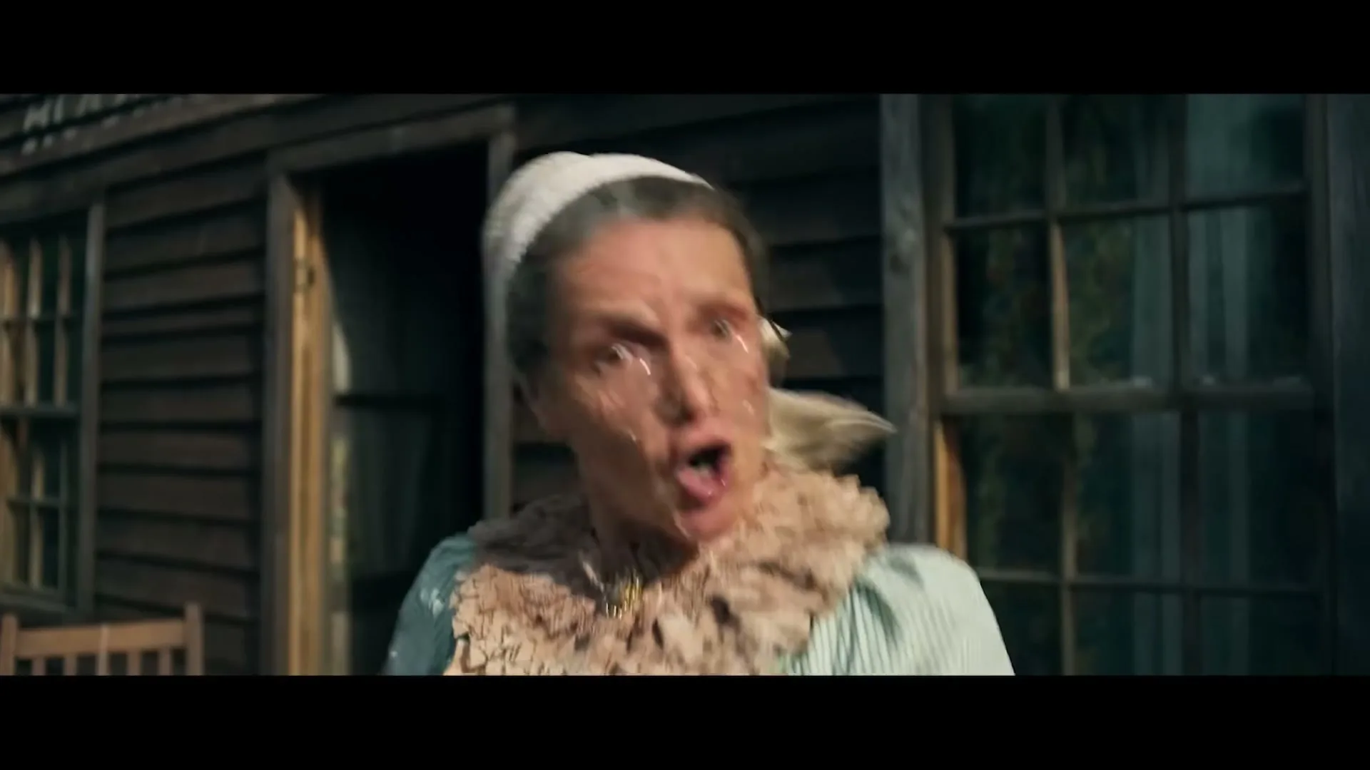 An older woman in a bonnet and glasses, wearing a ruffled collar, reacts with surprise and a splash of liquid on her face outside a rustic wooden house.