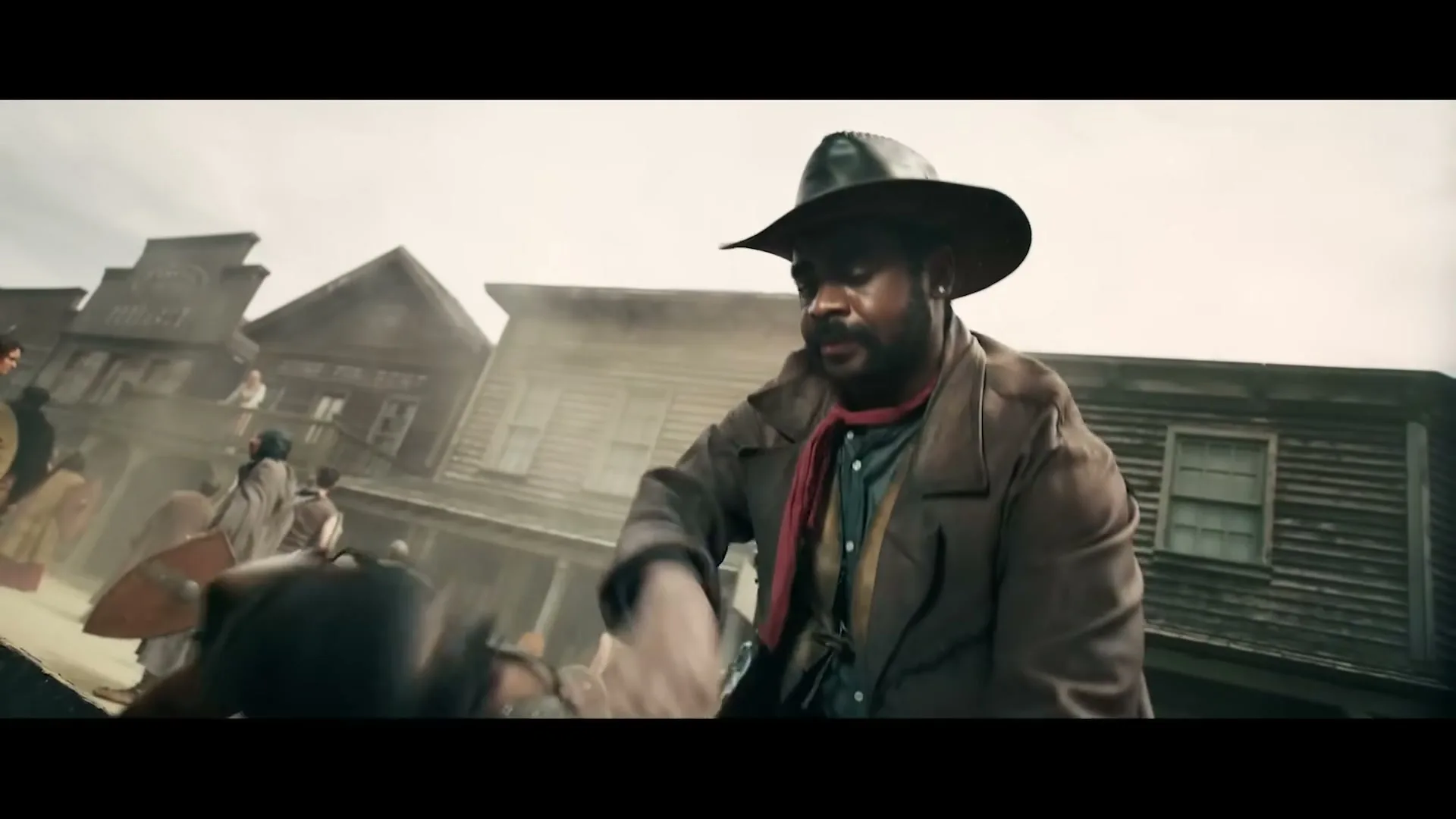 A cowboy in a brown coat and hat helps a blindfolded man with a head injury in a dusty old western town.