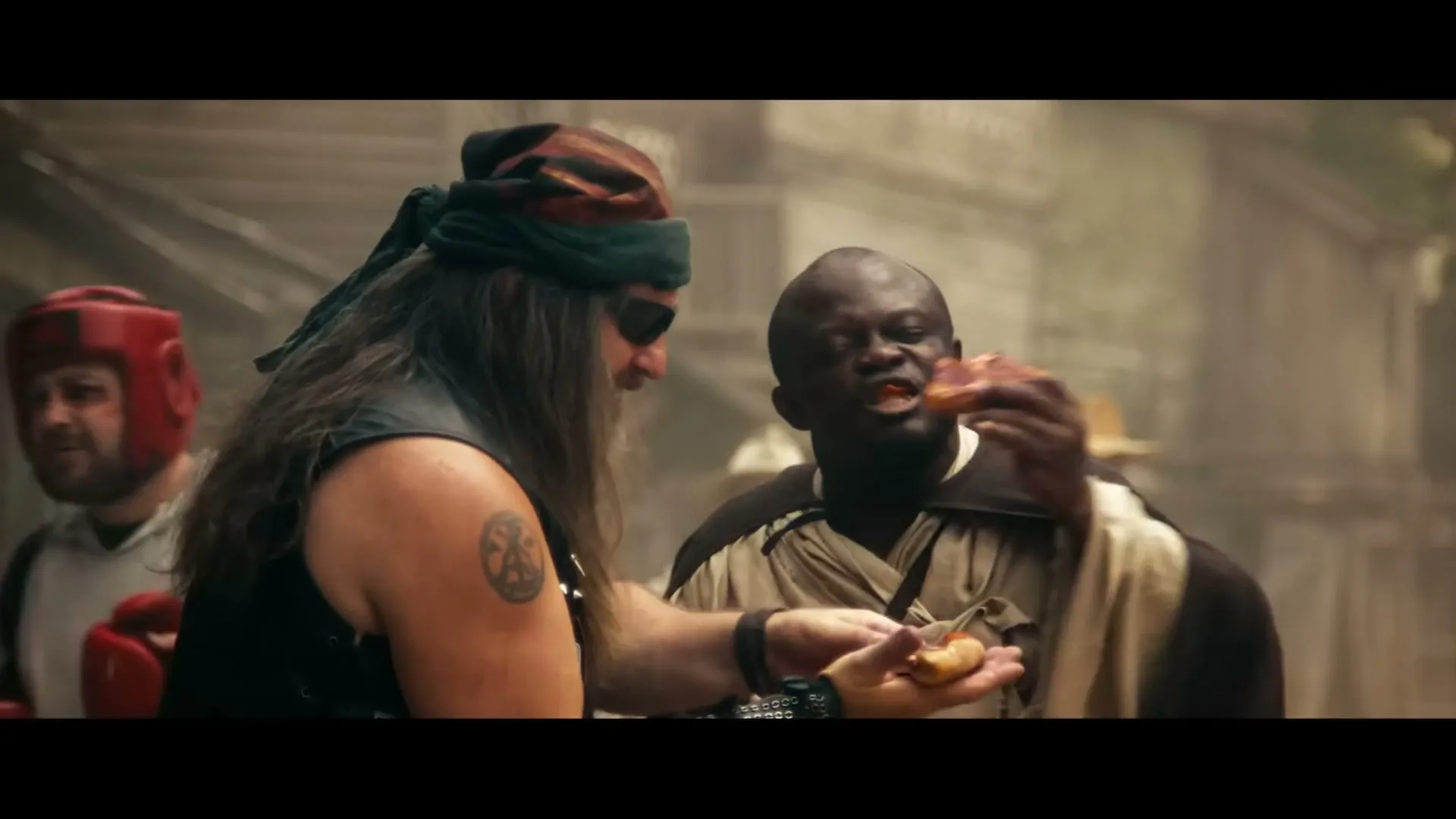 A man with long hair and a bandana offers a piece of pizza to another man in a robe, who is already eating a slice, in a dusty outdoor setting.