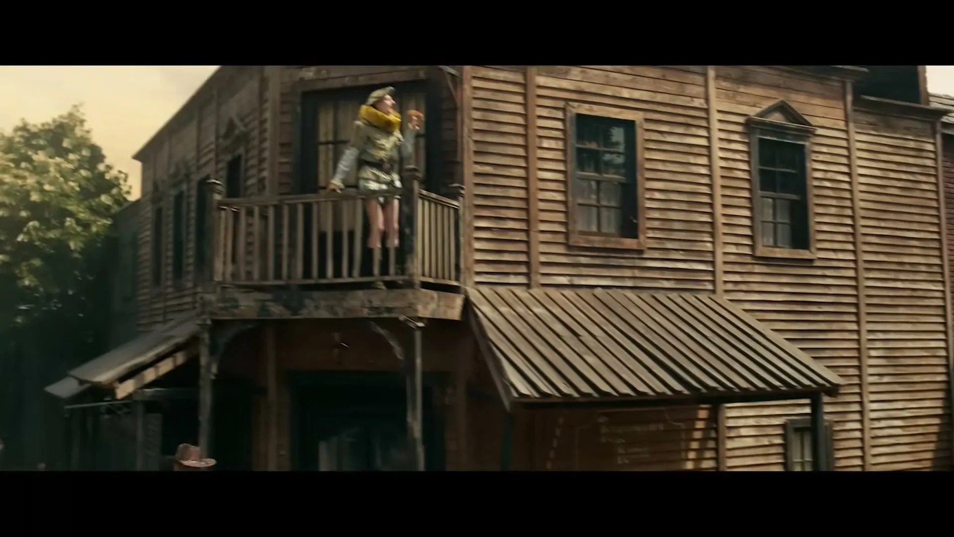 A woman in a yellow scarf and white shirt stands on a wooden balcony of an old western building, looking out over the town.