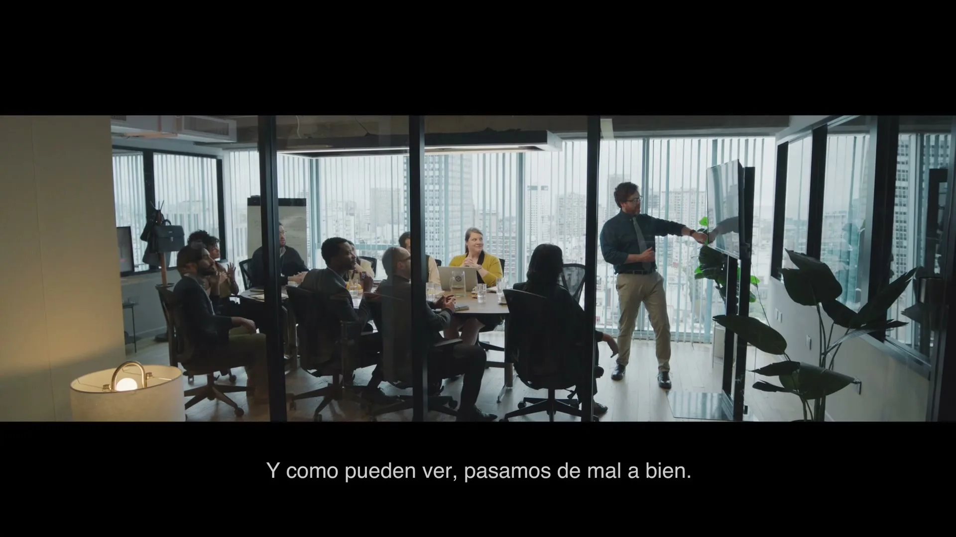 A diverse group of business people sit around a conference table while a male presenter points at a screen in a modern office.