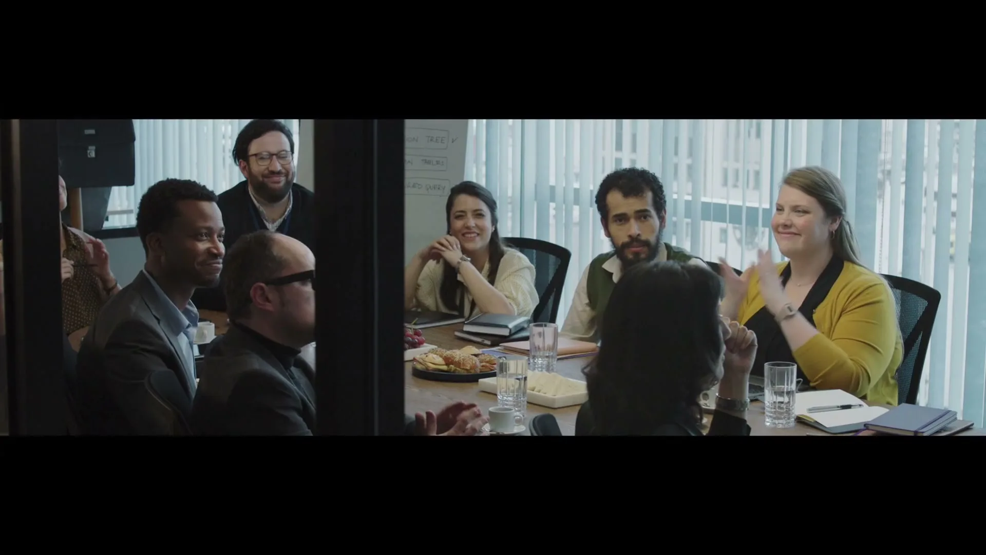 A diverse group of professionals sits around a conference table, smiling and clapping, in a modern office setting during the day.